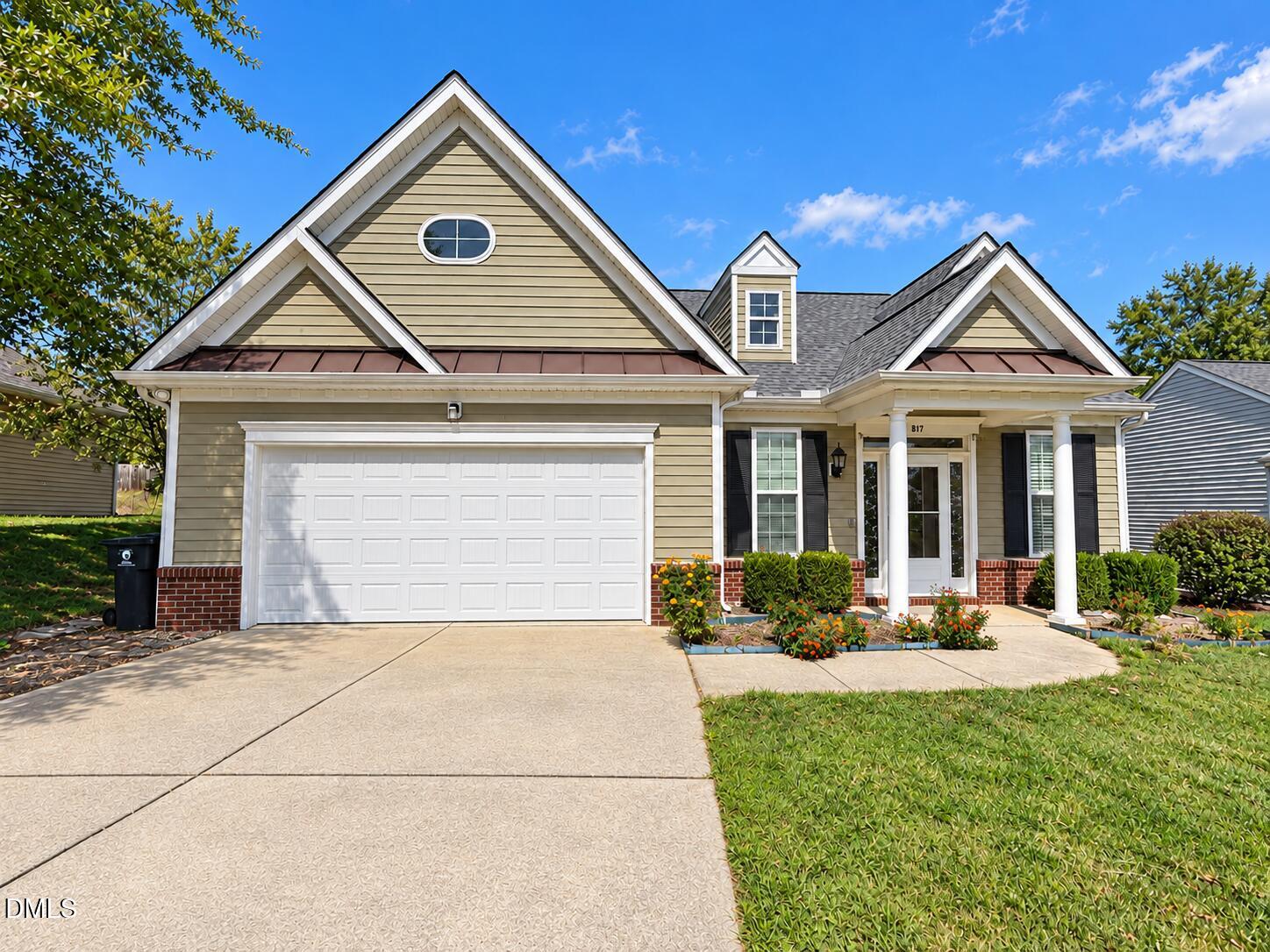 Beige gabled home with a two-car garage, columned entry, and landscaped flower beds.