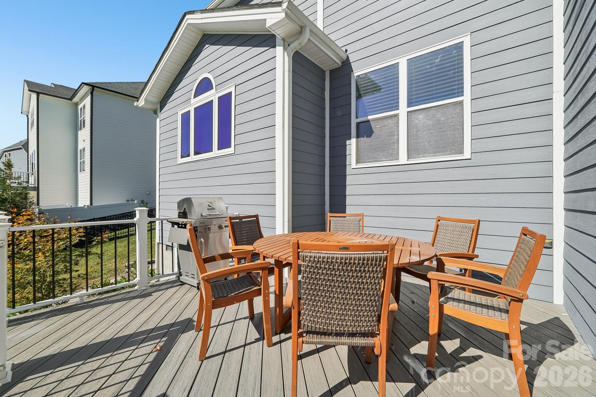 Furnished deck with a round wooden dining table and chairs, a stainless-steel grill, and gray house siding.