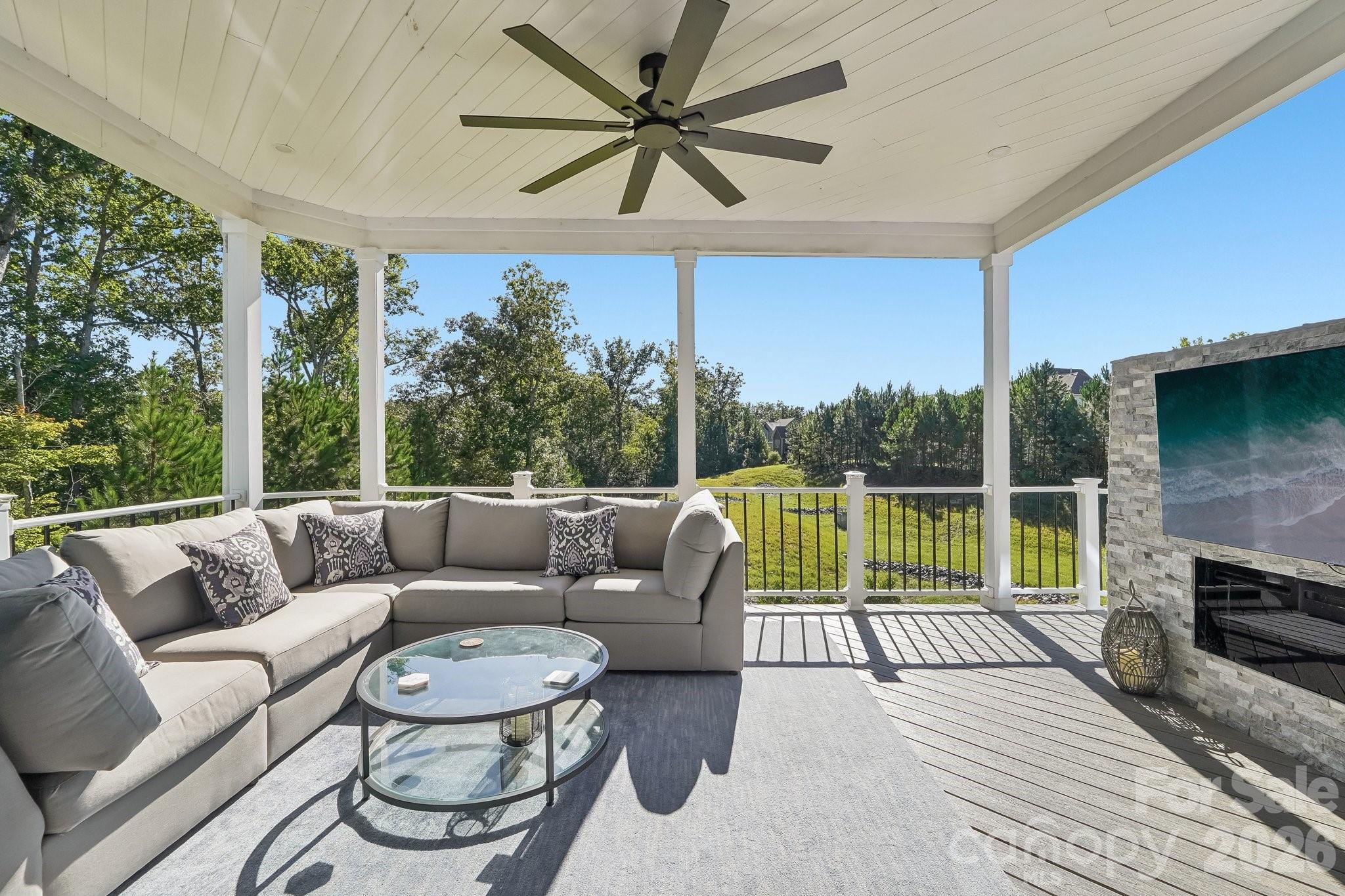 Covered patio with gray sectional seating, a ceiling fan, and stone outdoor fireplace overlooking a tree-lined lawn.