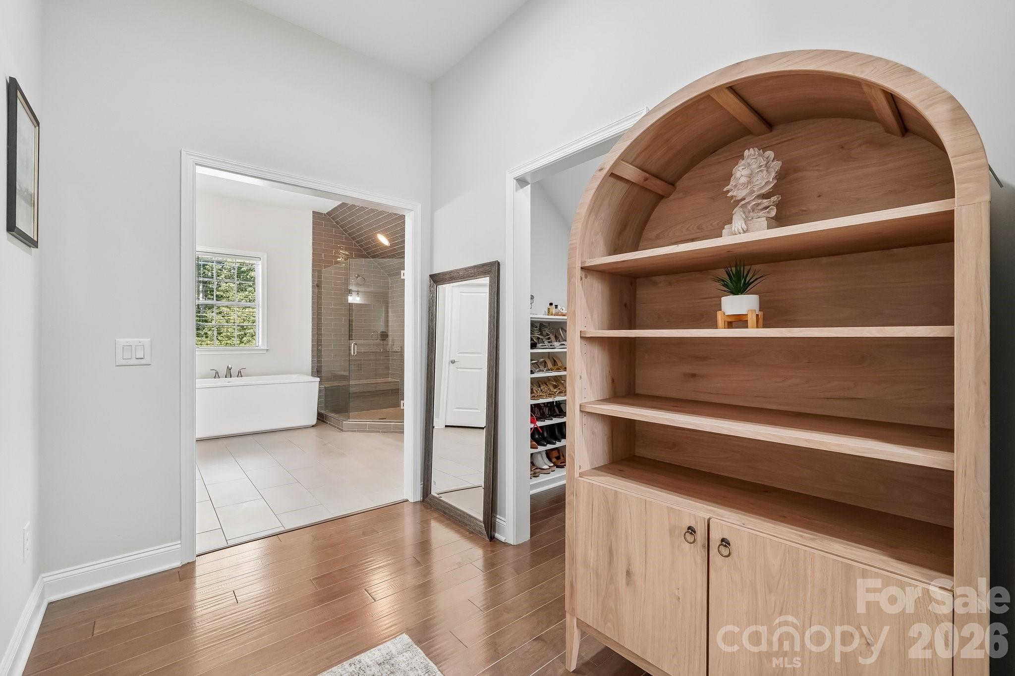 Arched light-wood built-in shelves beside a doorway to a tiled bathroom with a tub and glass shower.