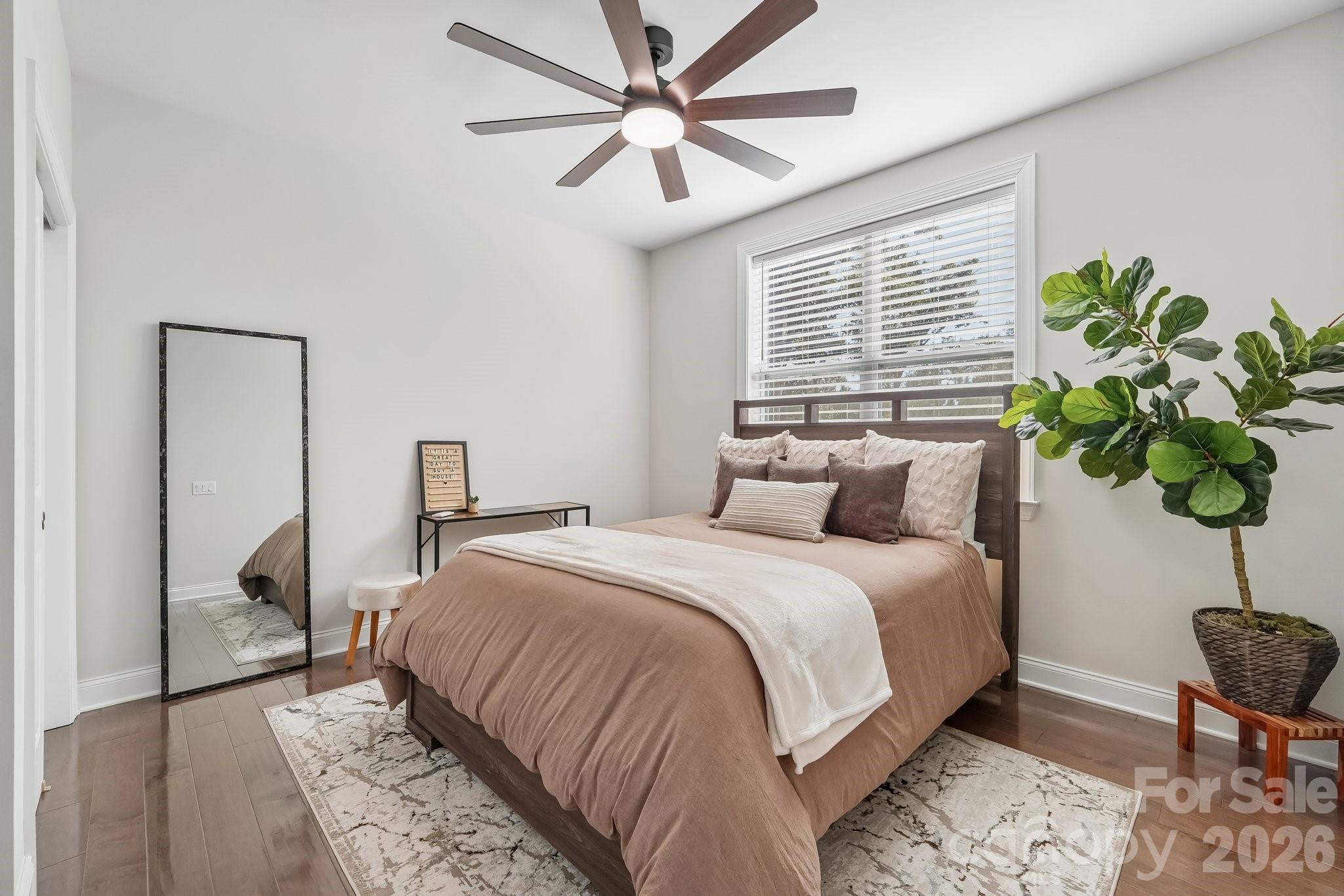 Staged bedroom with taupe bedding, a dark wood bed, ceiling fan, and large potted plant.