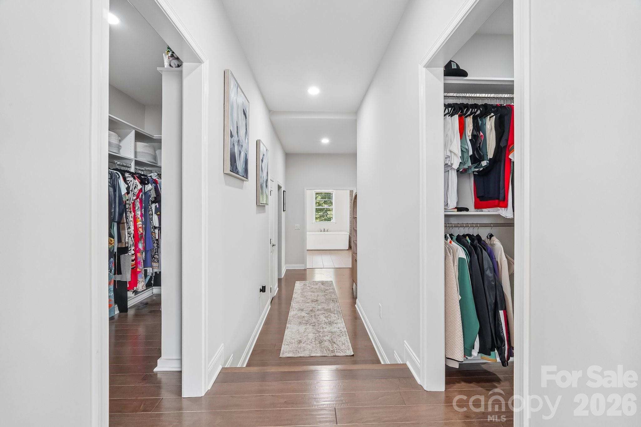Hallway with open, clothing-filled closets leading to a bathroom with a tub.