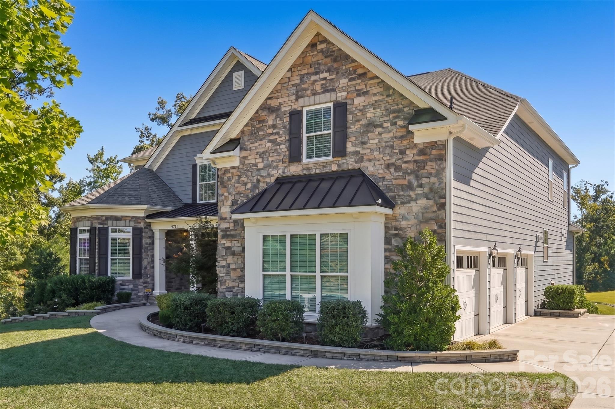 Two-story stone-front home with gray siding, gabled roof, three-car garage, and landscaped front yard.