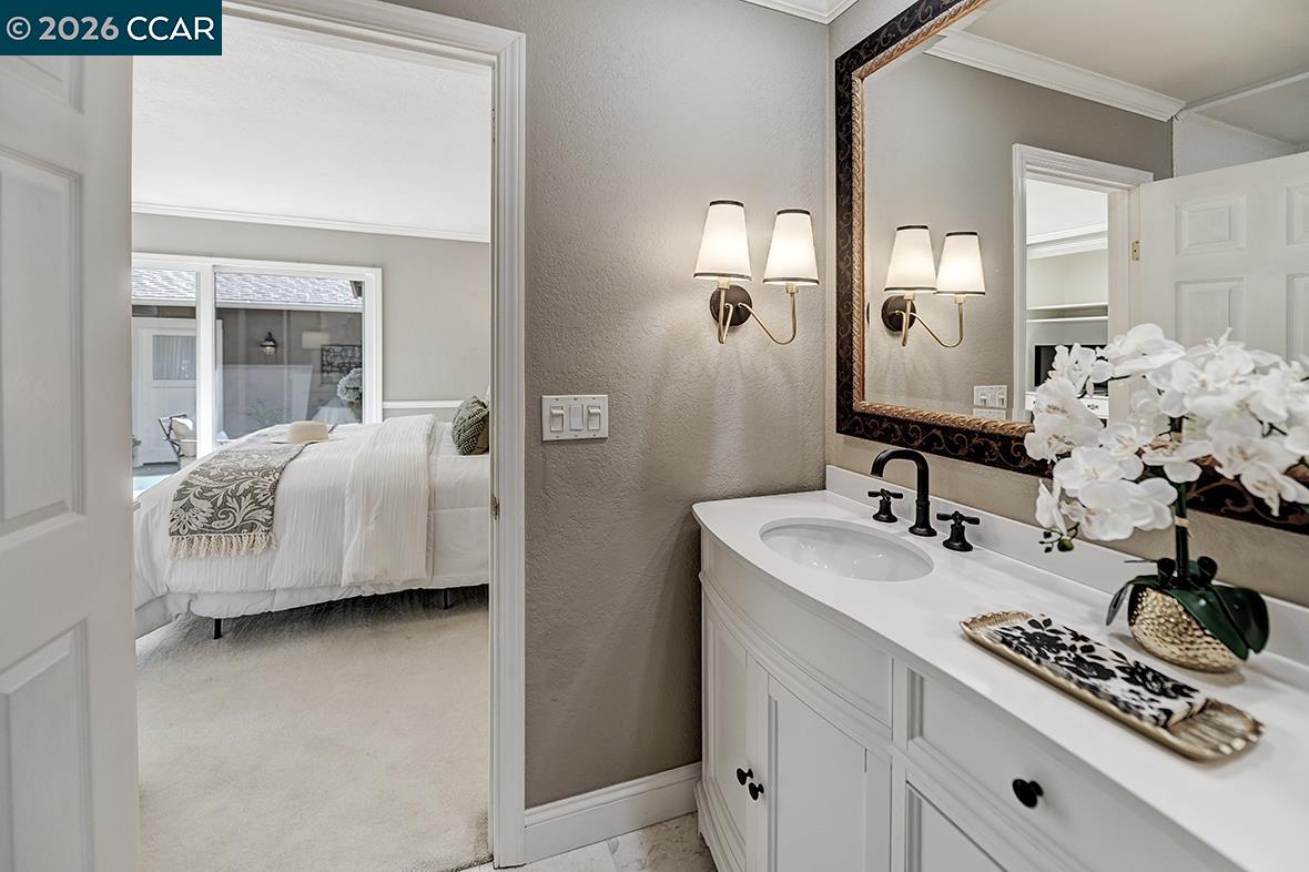 Bathroom vanity with an ornate mirror and flowers, looking through a doorway into a staged bedroom.