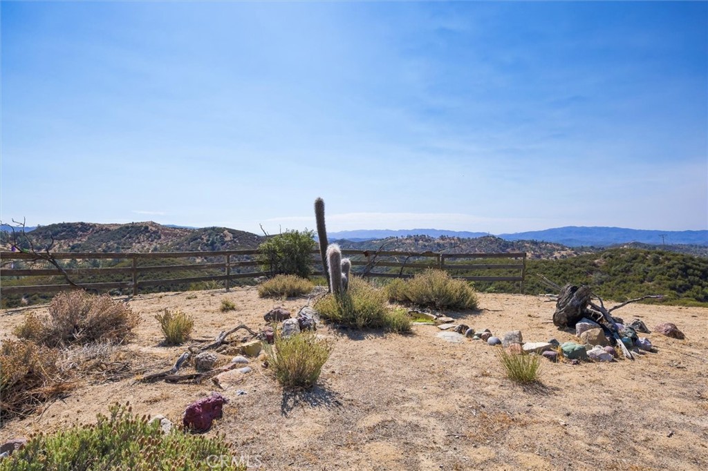 Dry rural property yard with cacti, a wooden rail fence, and distant hills.