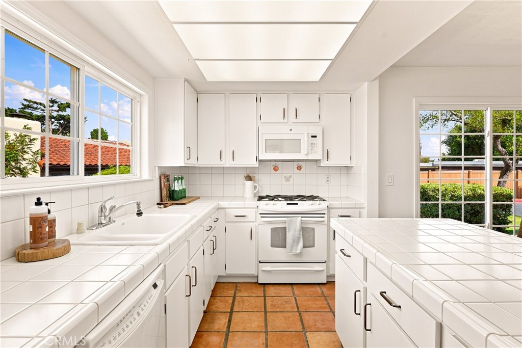 Bright white kitchen with tiled counters, terracotta floors, and large windows overlooking the yard.