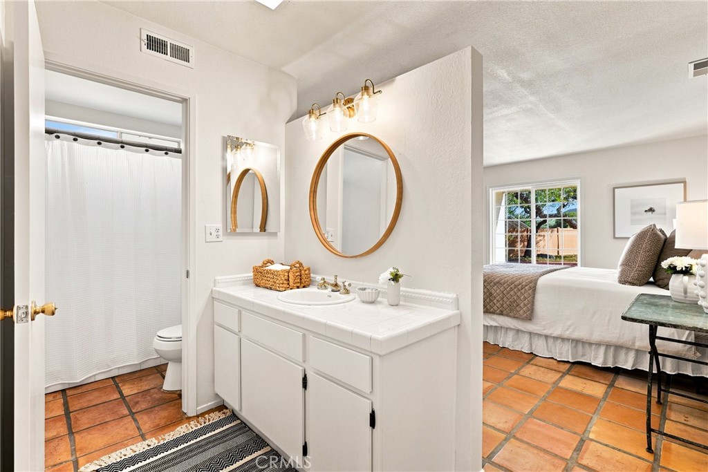 Bathroom with a white vanity and oval mirror, opening onto a bedroom with terracotta tile flooring.