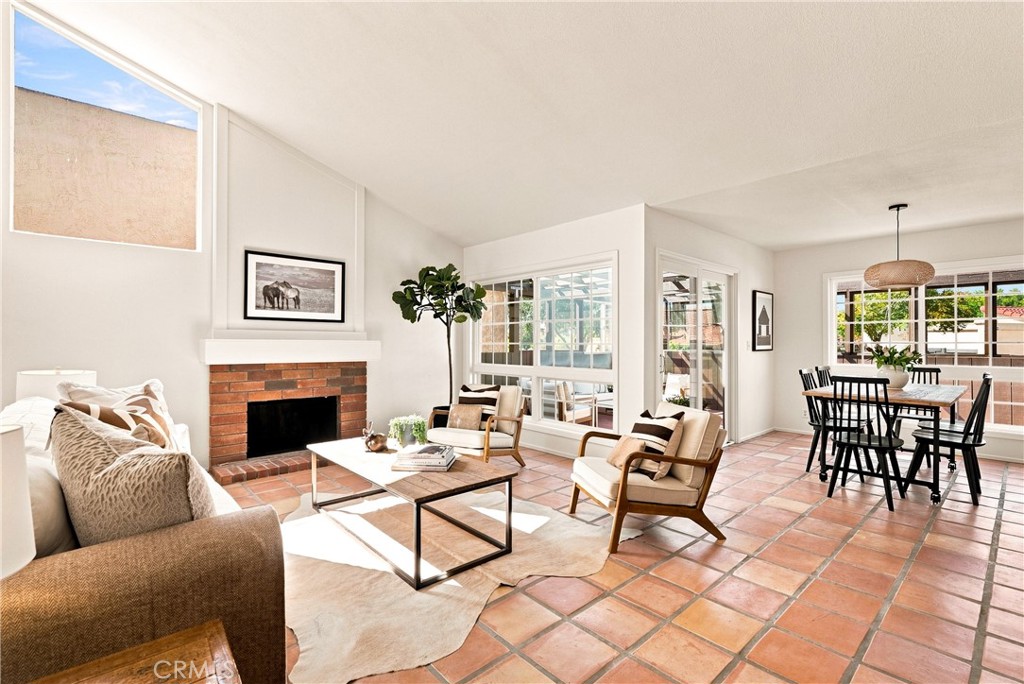 Open-plan living and dining room with terracotta tile floors, brick fireplace, and large windows.