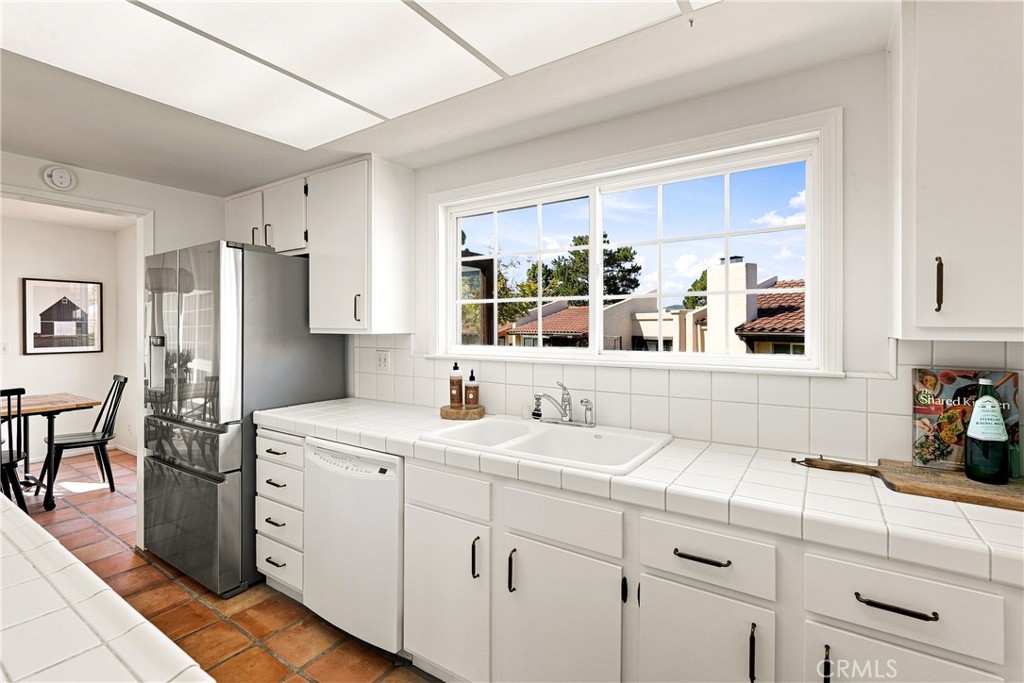 White kitchen with tiled counters, stainless steel refrigerator, and a sink beneath a large window.