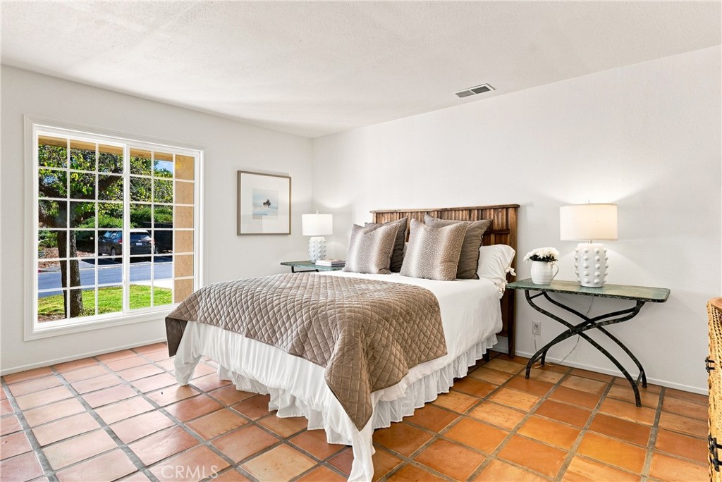 Staged bedroom with a wooden headboard, terracotta tile floor, and large window overlooking greenery.