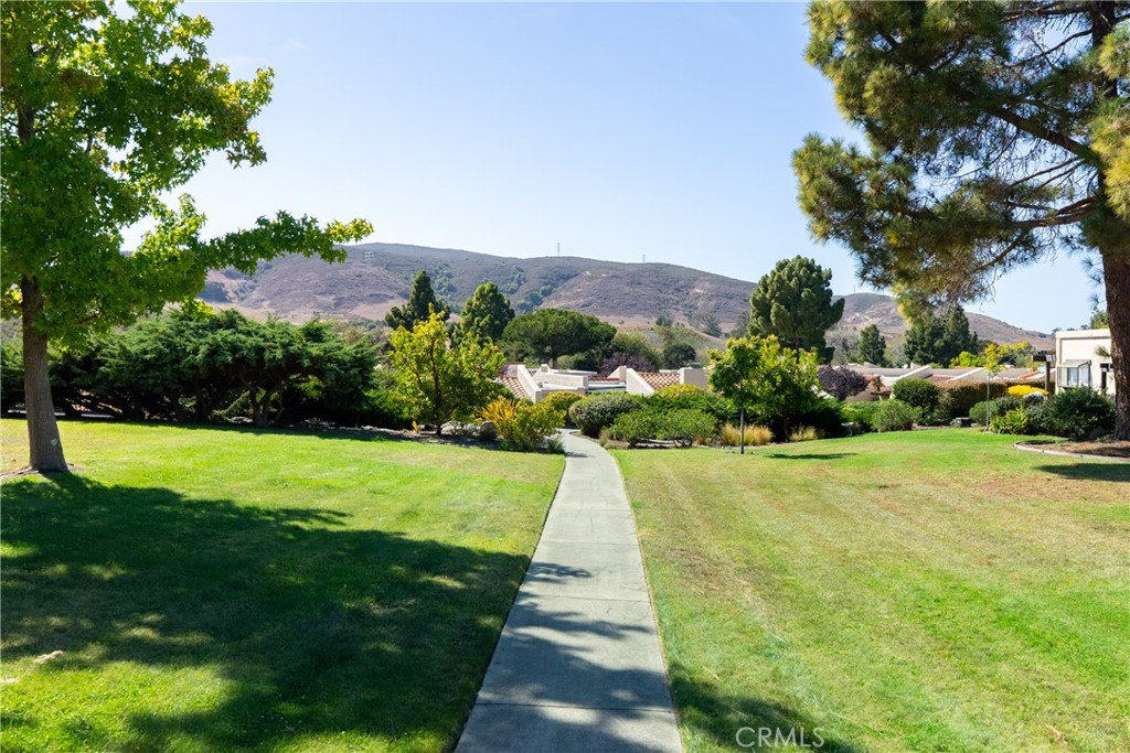Landscaped community grounds with a curving sidewalk, lawns, mature trees, and hills beyond.