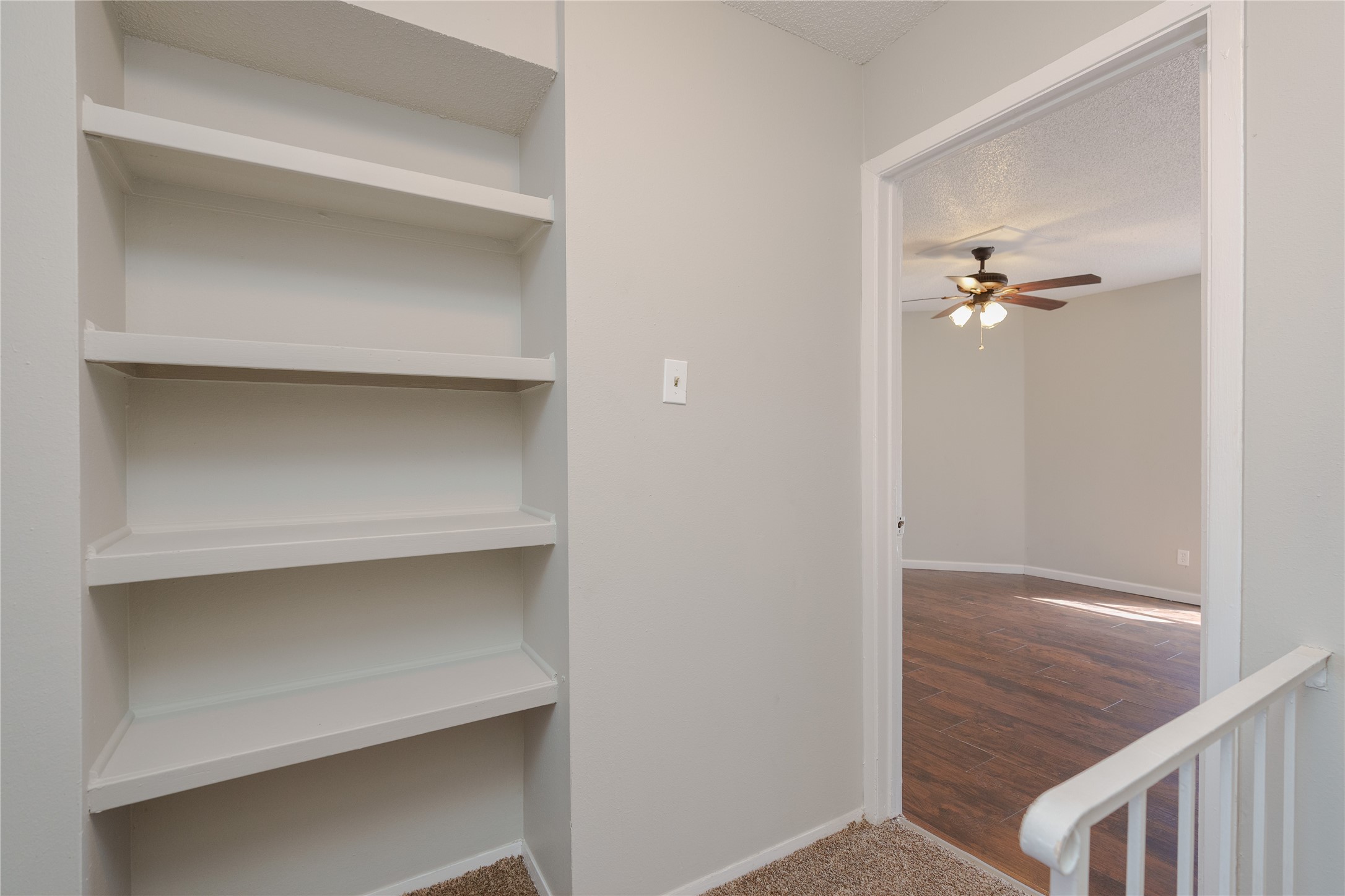 Built-in white shelves beside a carpeted landing, overlooking an empty room with dark wood floors and a ceiling fan.