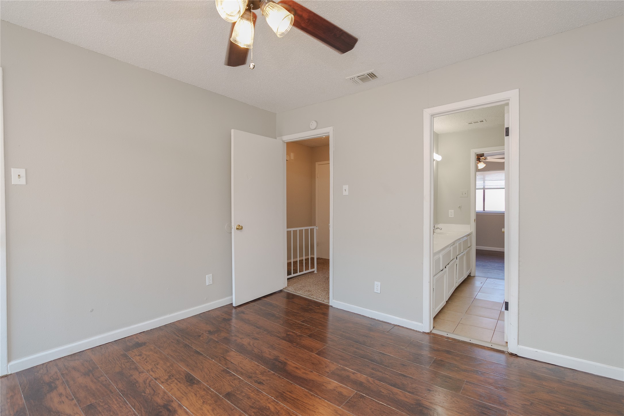 Empty room with dark plank flooring and ceiling fan, opening to a hall and bathroom.