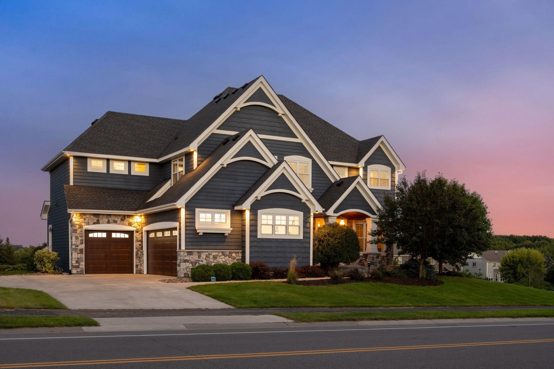 Two-story dark blue-gray house with white gables, stone accents, and two garage doors at dusk.
