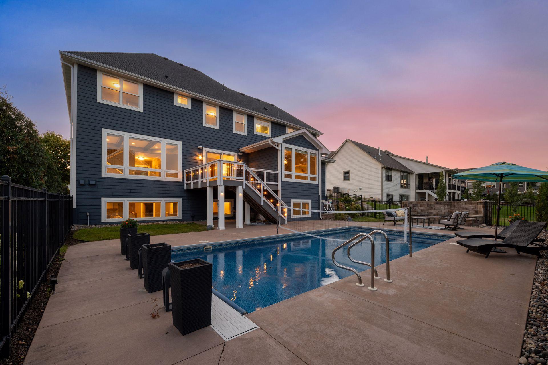 Rear of a dark blue home with a raised deck and illuminated poolside patio at dusk.