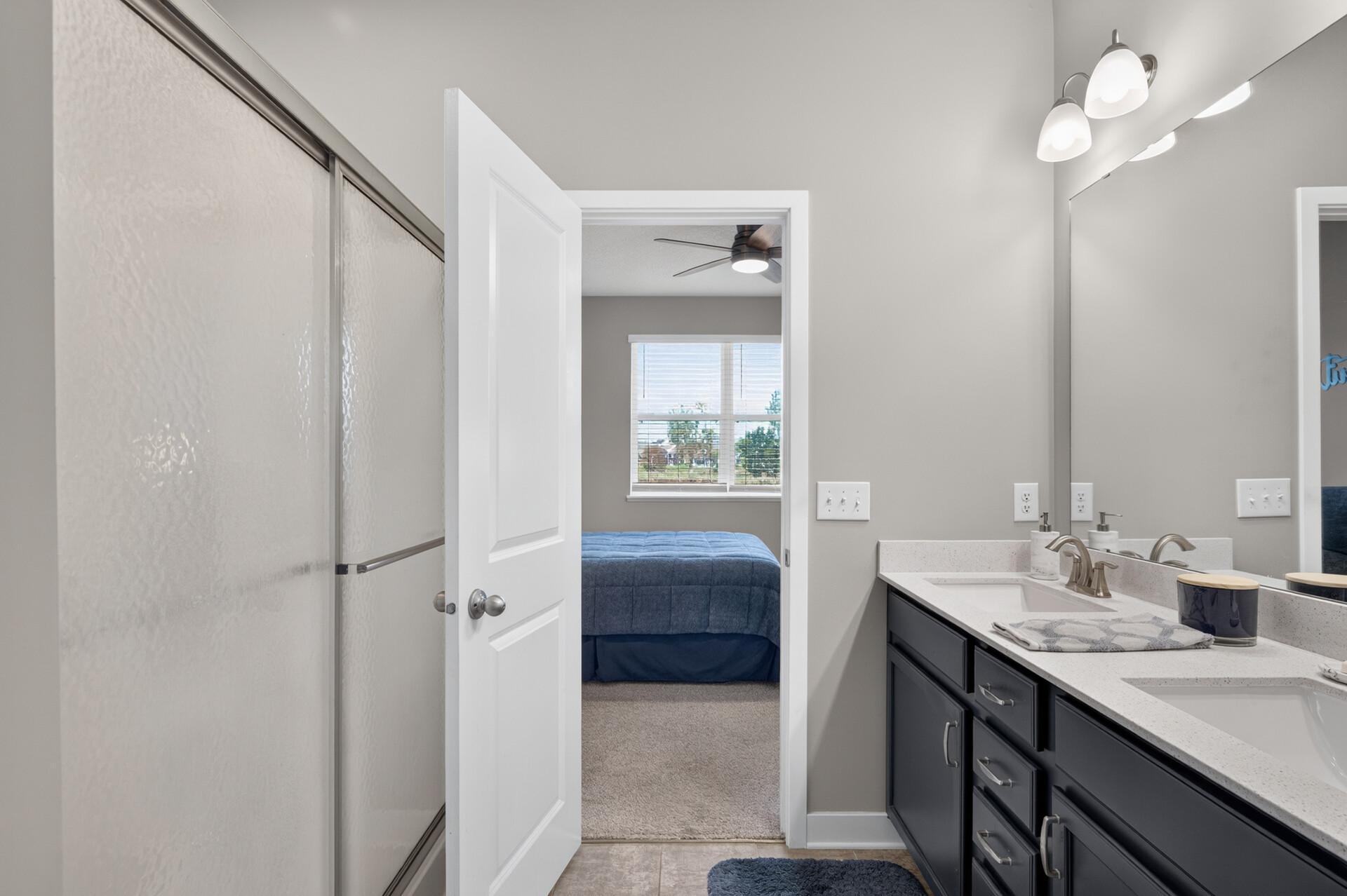 Bathroom with glass shower and double vanity, looking through an open door toward a bedroom with a blue bed.