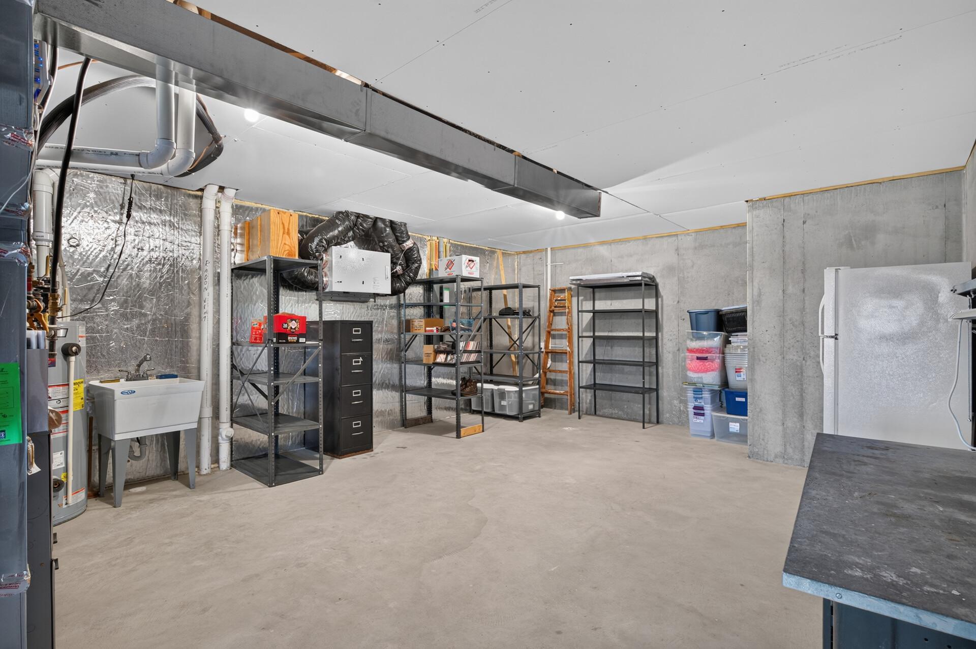 Unfinished basement utility room with exposed ductwork, metal storage shelves, a utility sink, and a refrigerator.