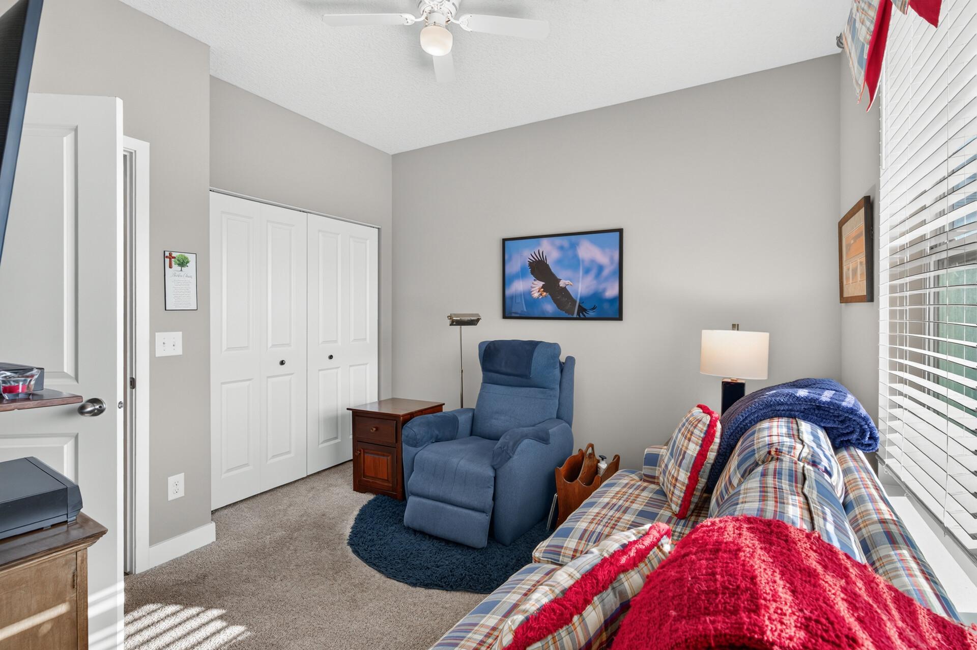 Furnished bedroom with a plaid bed, blue recliner, white closet doors, and bright window.
