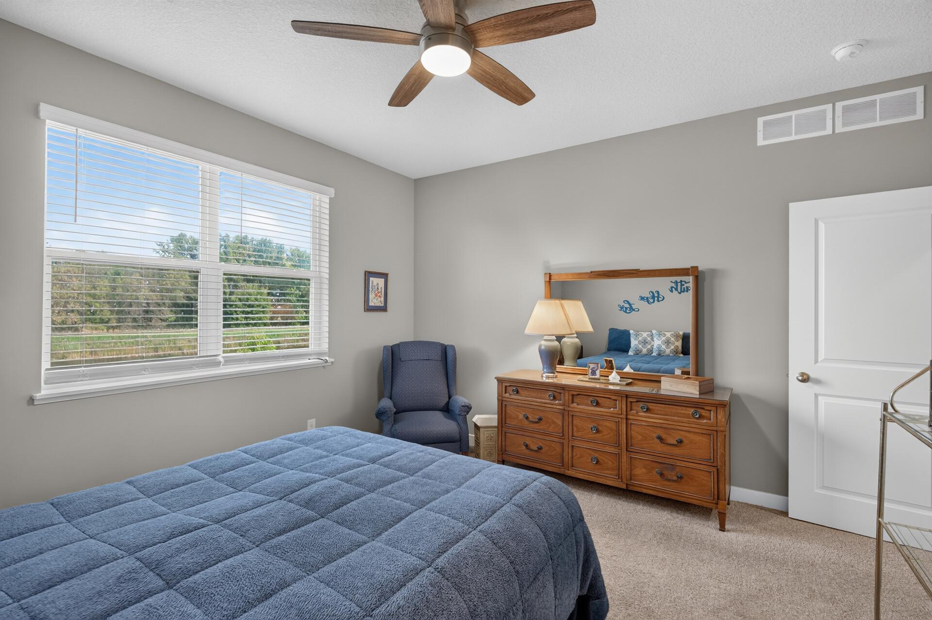 Bedroom with a blue quilted bed, wooden dresser and mirror, and large window with white blinds.