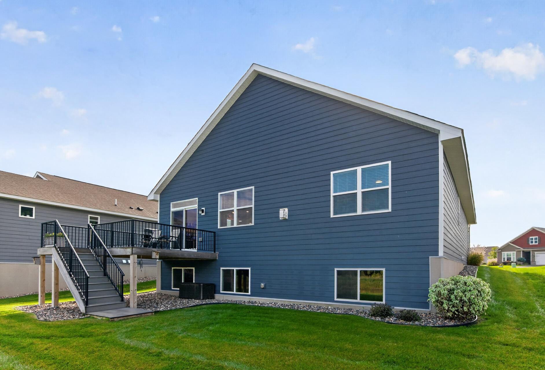 Blue-sided home’s rear exterior with an elevated deck, black railings, and a broad green lawn.