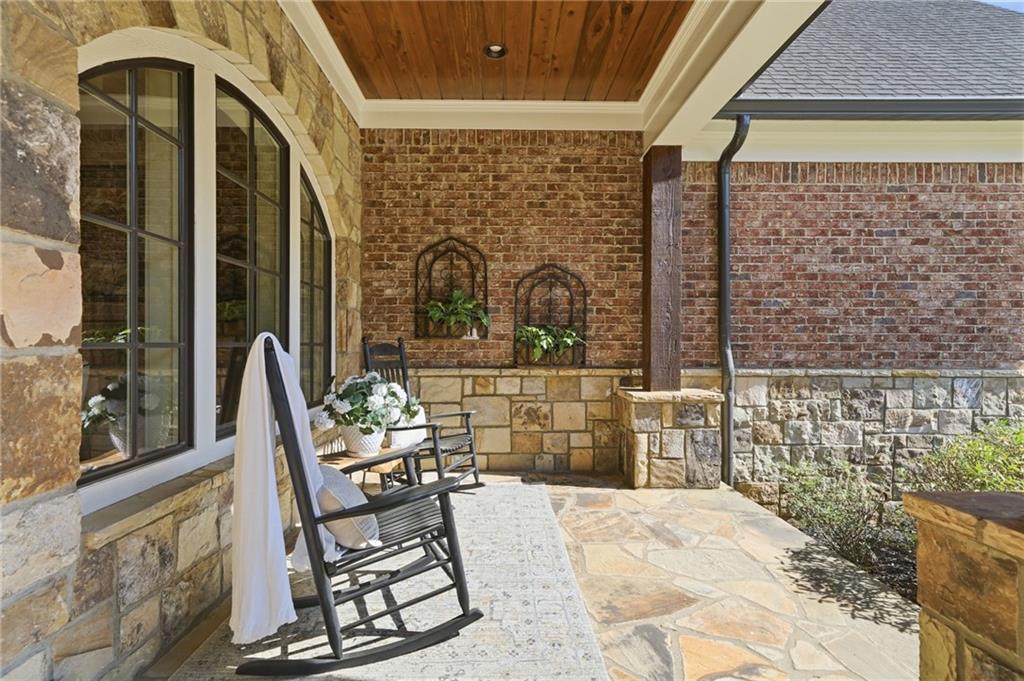 Covered stone patio with a rocking chair, arched windows, and a wood-plank ceiling.