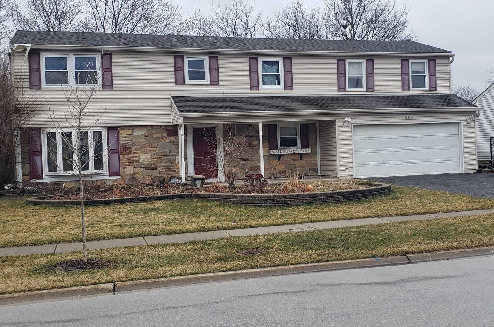 Two-story beige home with stone facade, burgundy shutters, and an attached garage.