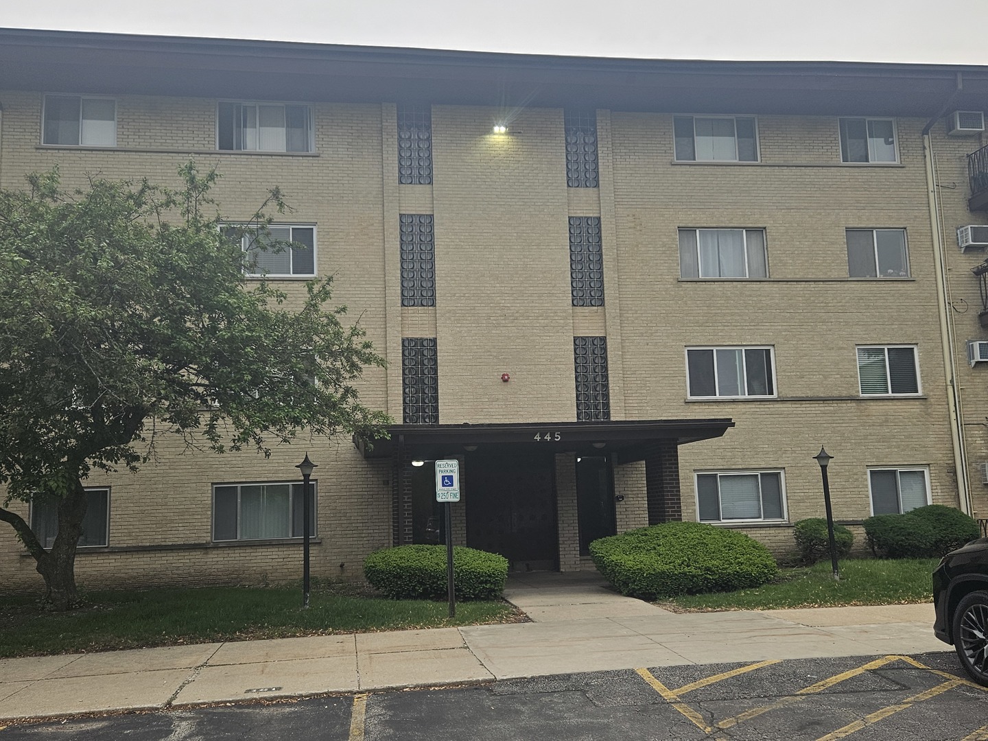 Tan brick apartment building at 445, with a recessed entrance, glass-block columns, and landscaped shrubs.