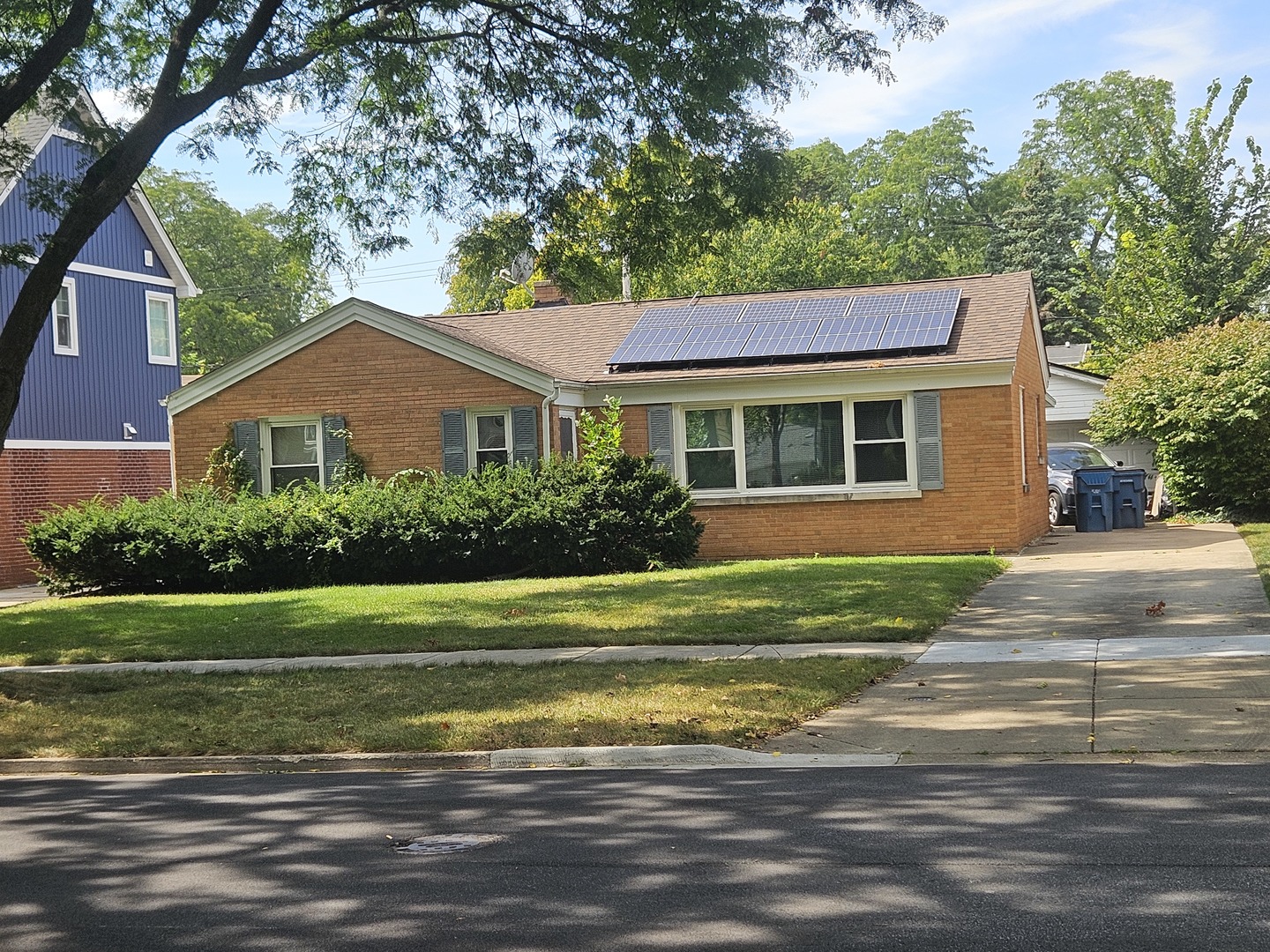 Red-brick ranch house with rooftop solar panels, front shrubs, and a driveway.