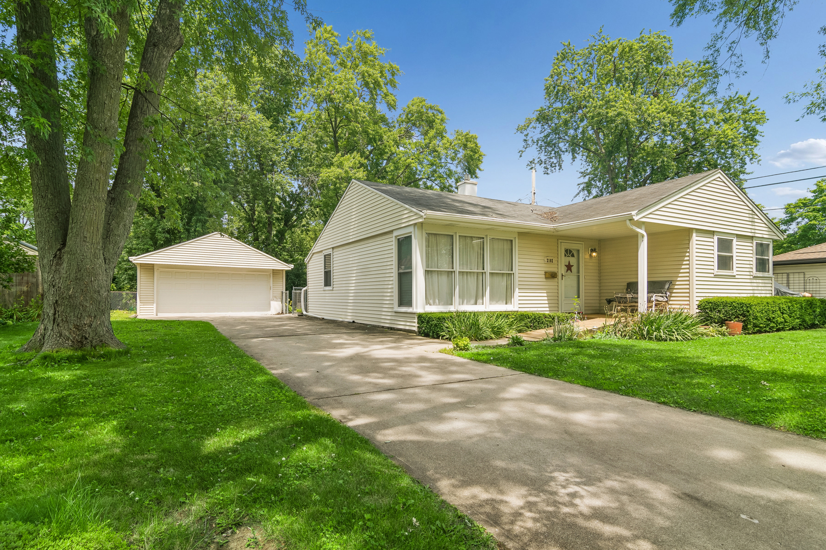 Single-story house with a detached garage, wide driveway, and mature trees.