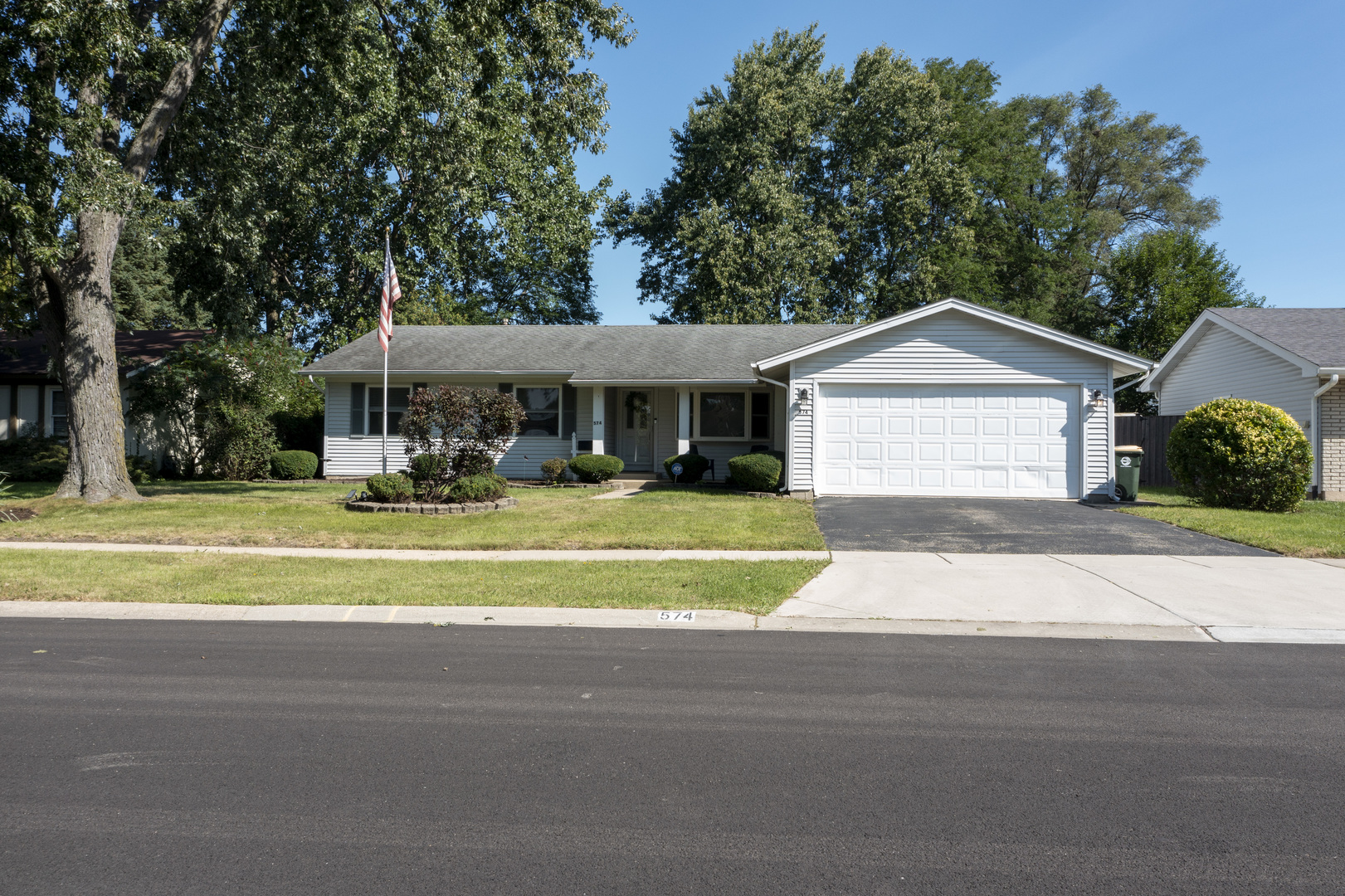 Single-story gray ranch home with a two-car garage, landscaped front lawn, and American flag.
