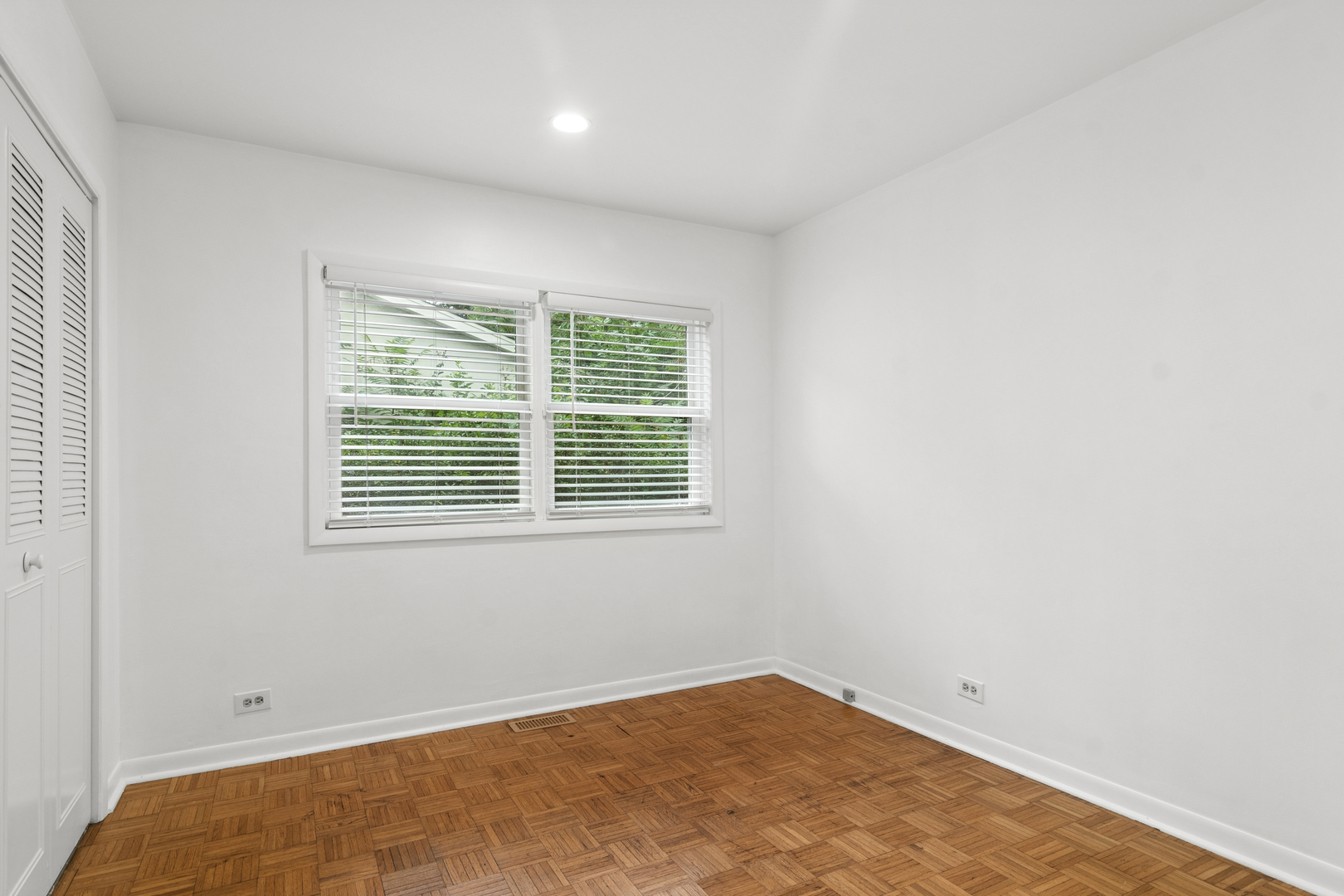 Empty room with parquet flooring, louvered closet doors, and two windows with white blinds.