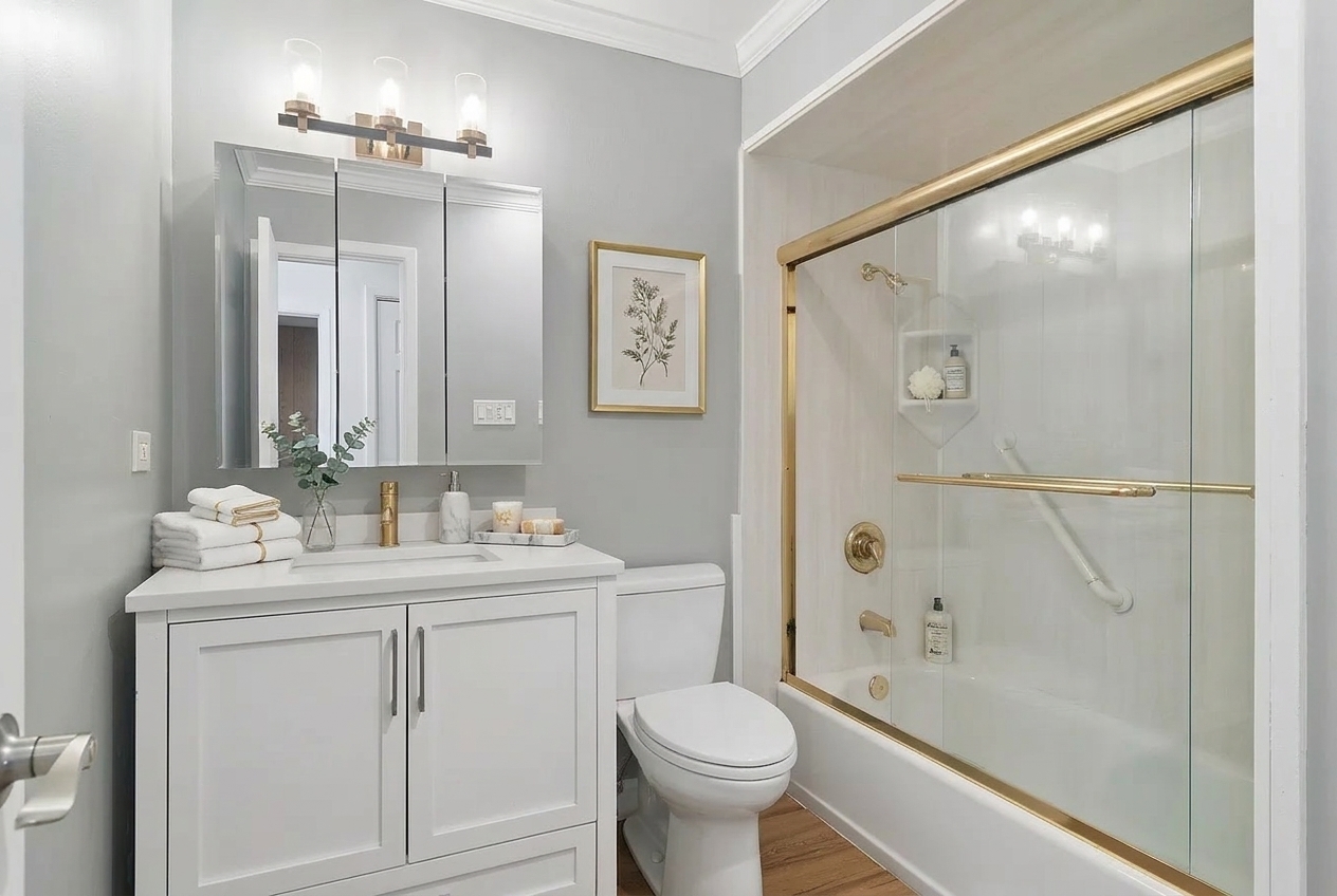 Renovated bathroom with a white vanity, toilet, and glass shower enclosure with gold trim.