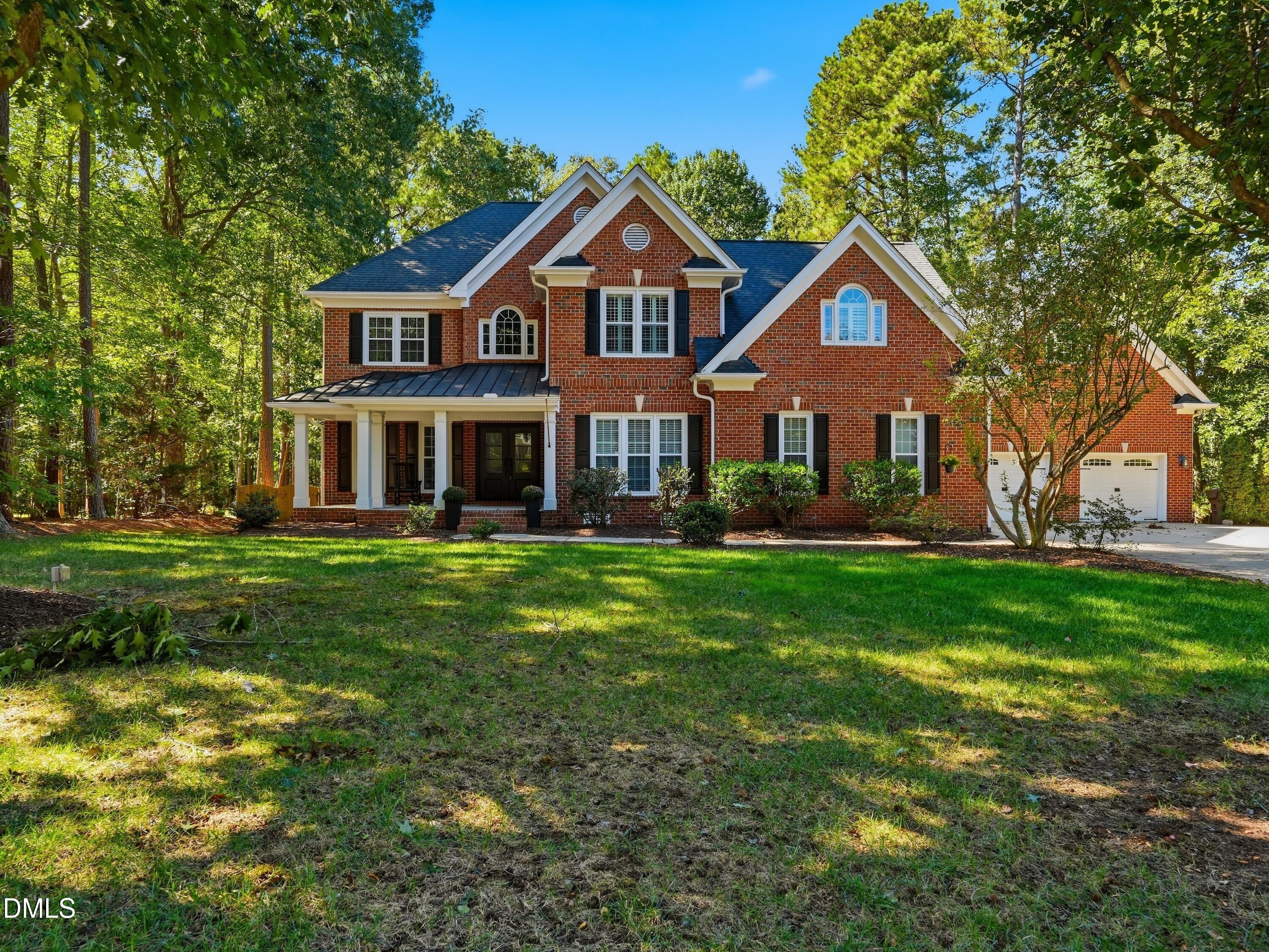 Two-story red-brick house with a covered porch and attached garage, surrounded by mature trees.