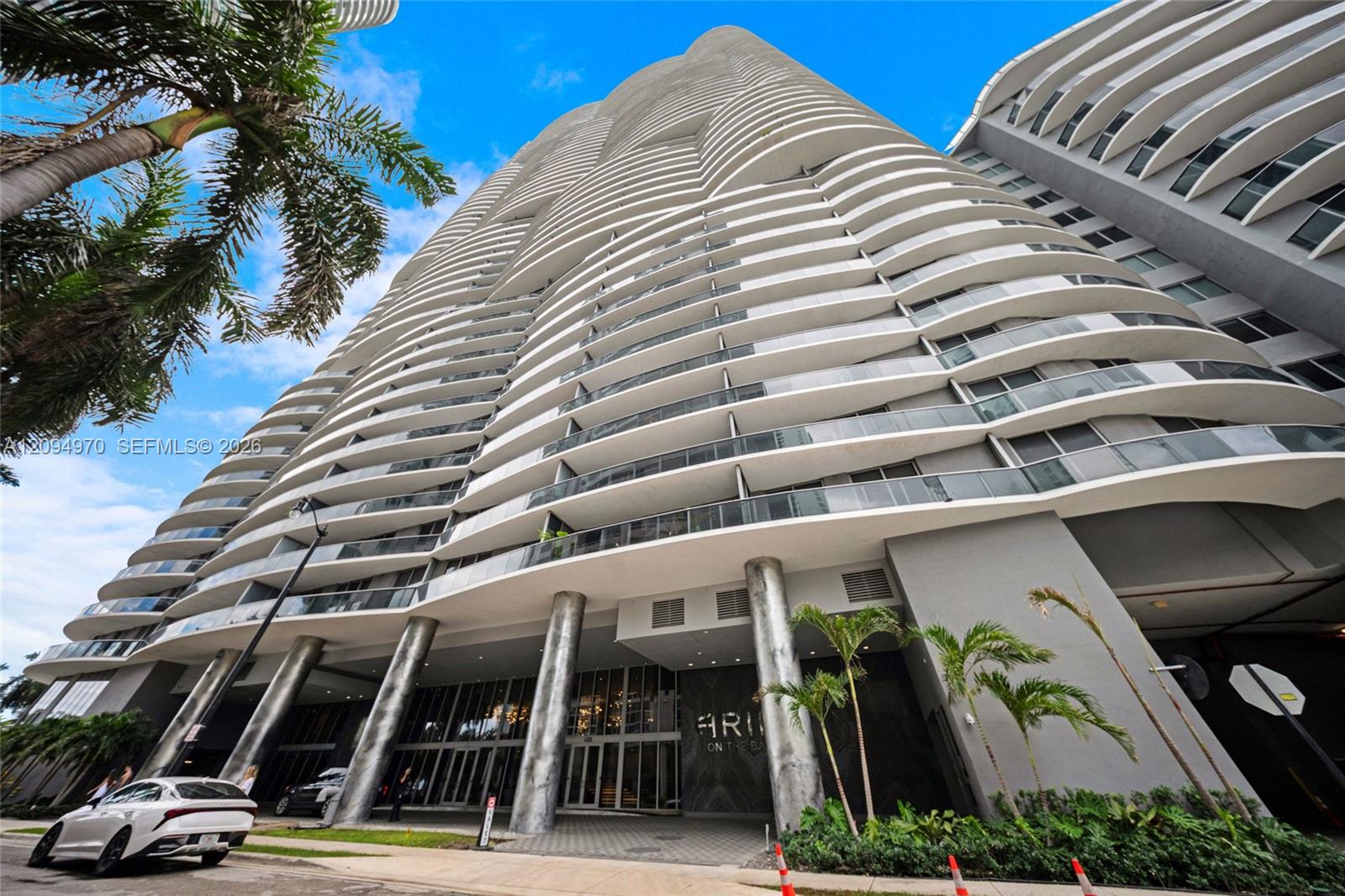 Modern condominium tower with sweeping curved balconies, palm trees, and a street-level entrance.