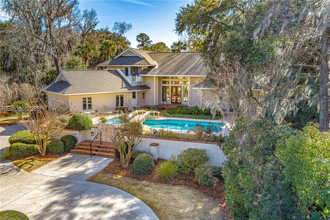 Beige home with a turquoise pool, curved driveway, and mature trees.