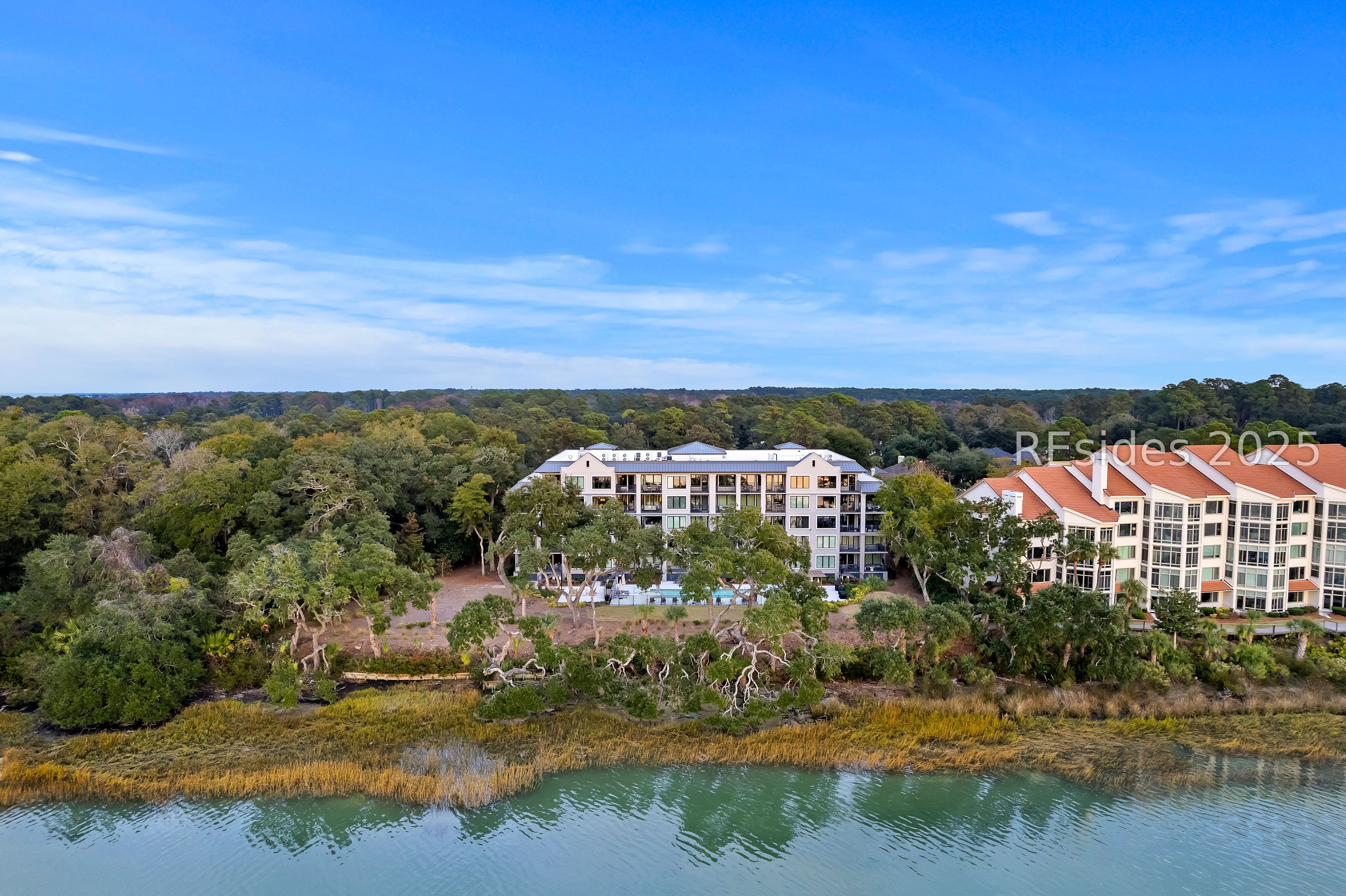 Aerial view of waterfront condominiums beside a tree-lined marsh, with a pool between the buildings.
