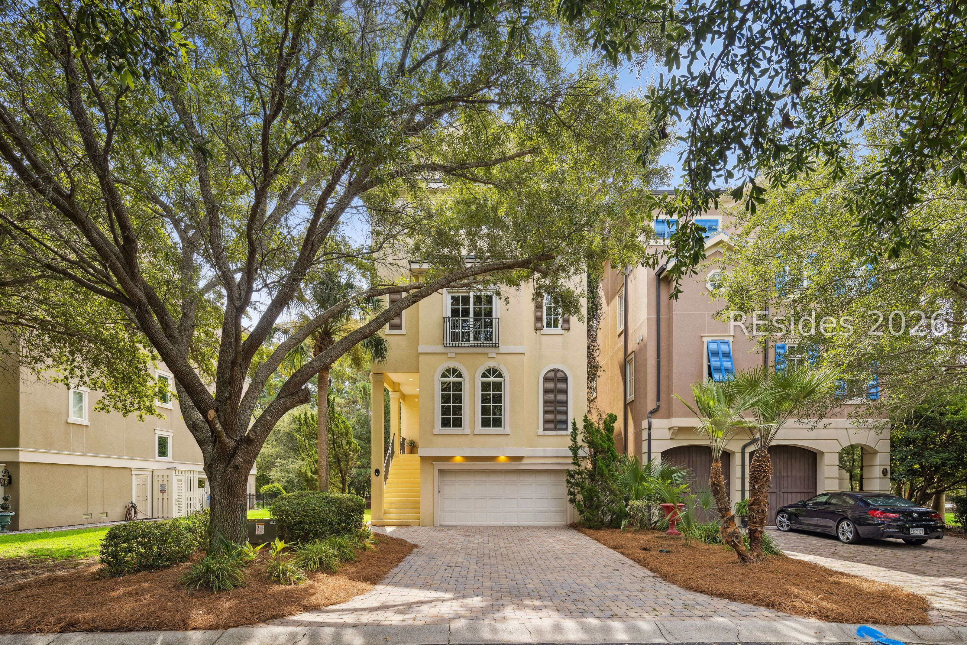 Stucco home with a paver driveway, garage, blue shutters, and a large shade tree.