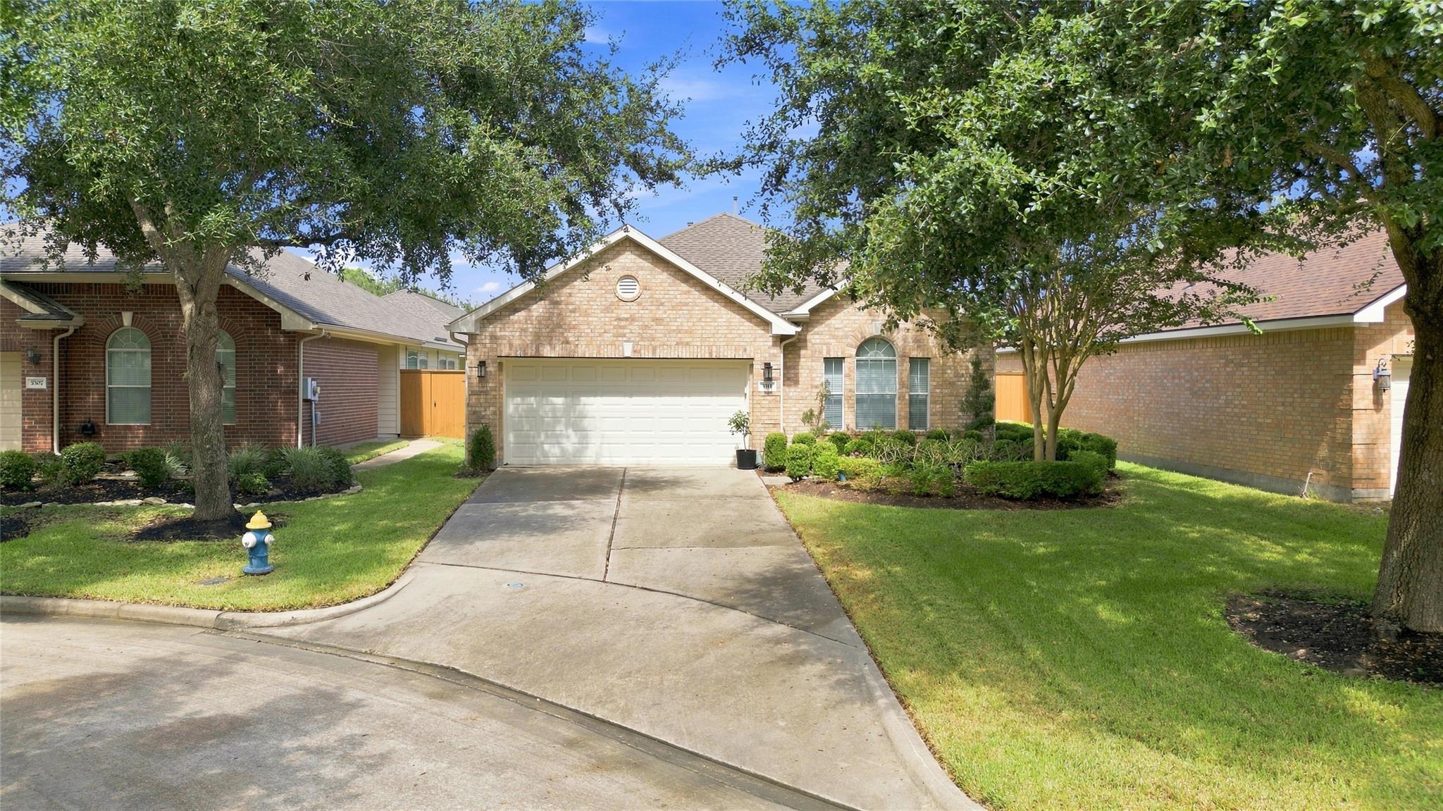 Tan brick single-story home with a two-car garage, curved driveway, and mature trees.