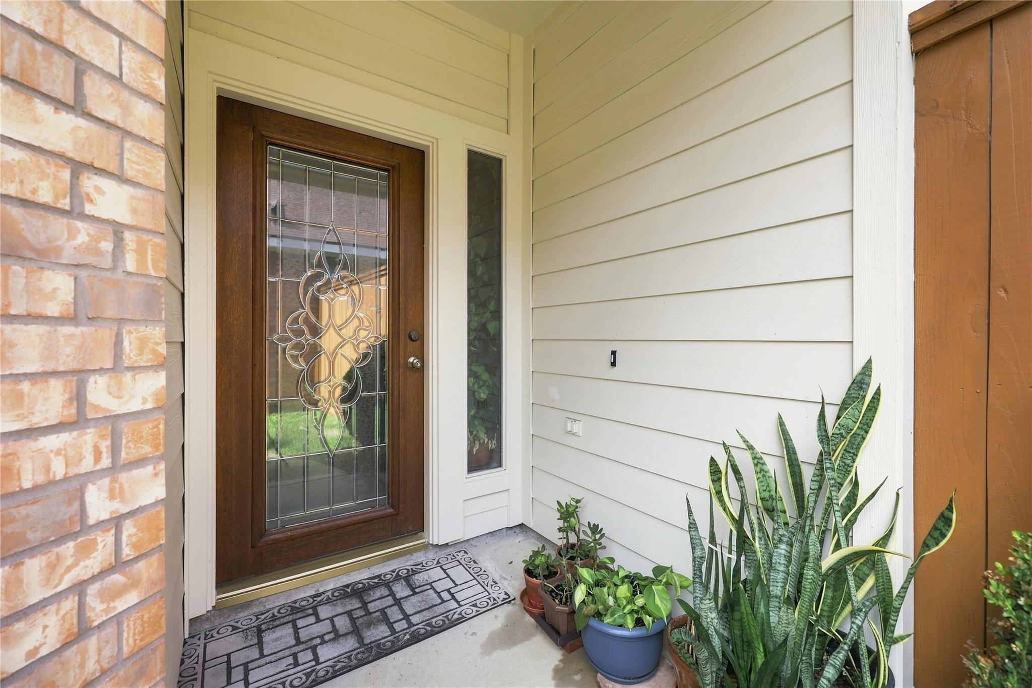 Covered entry with decorative glass front door, brick wall, potted plants, and patterned doormat.