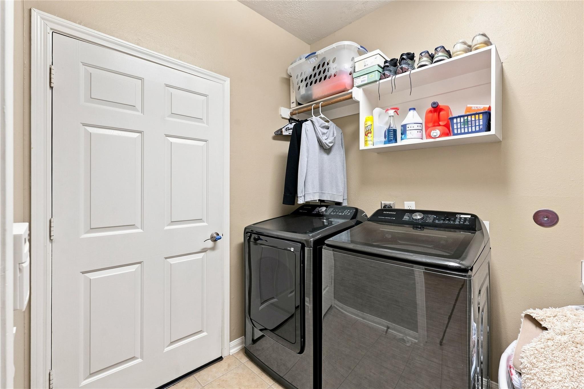 Laundry room with dark washer and dryer beneath shelves of shoes and laundry supplies.