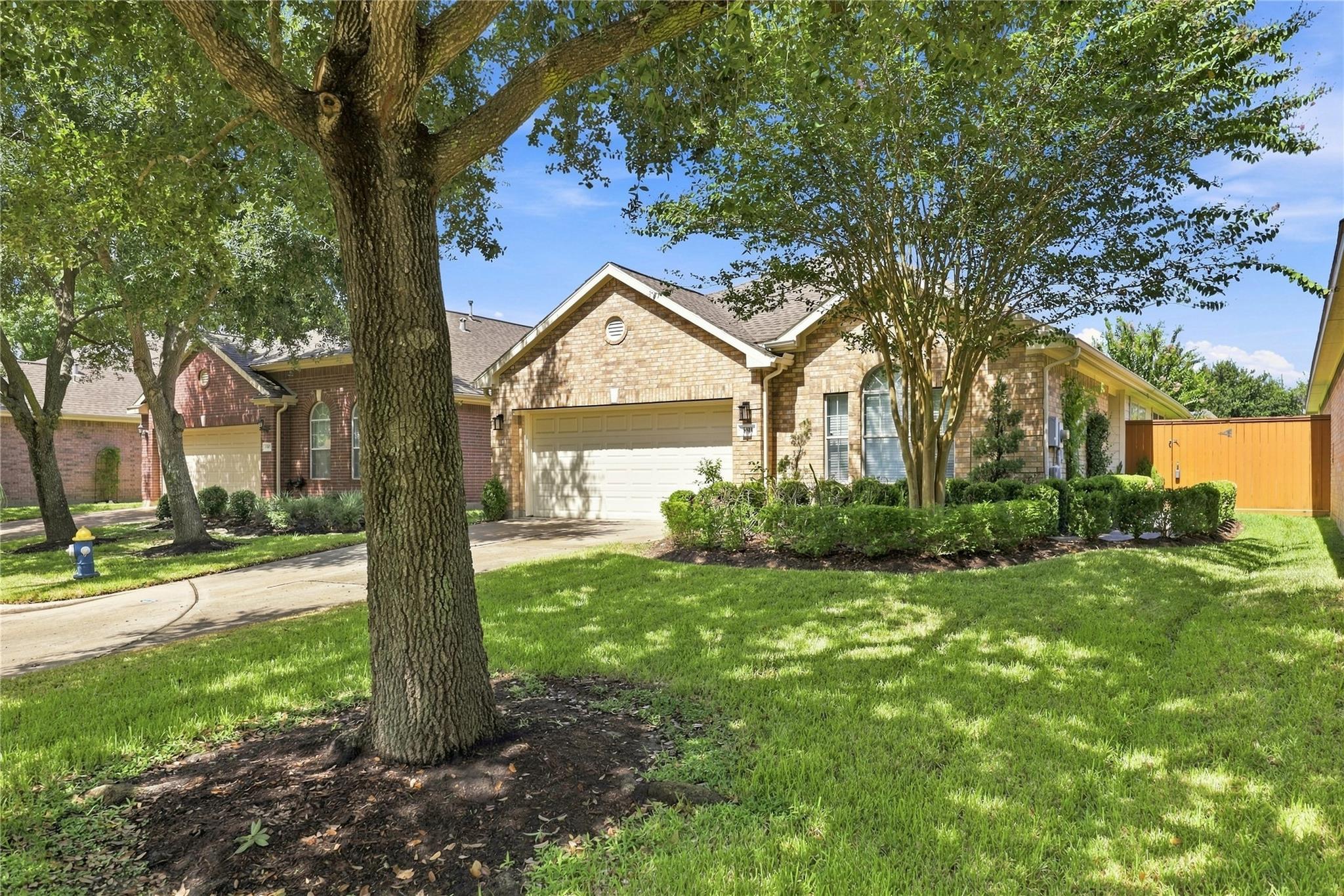 Brick home with two-car garage behind mature shade trees and a landscaped lawn.