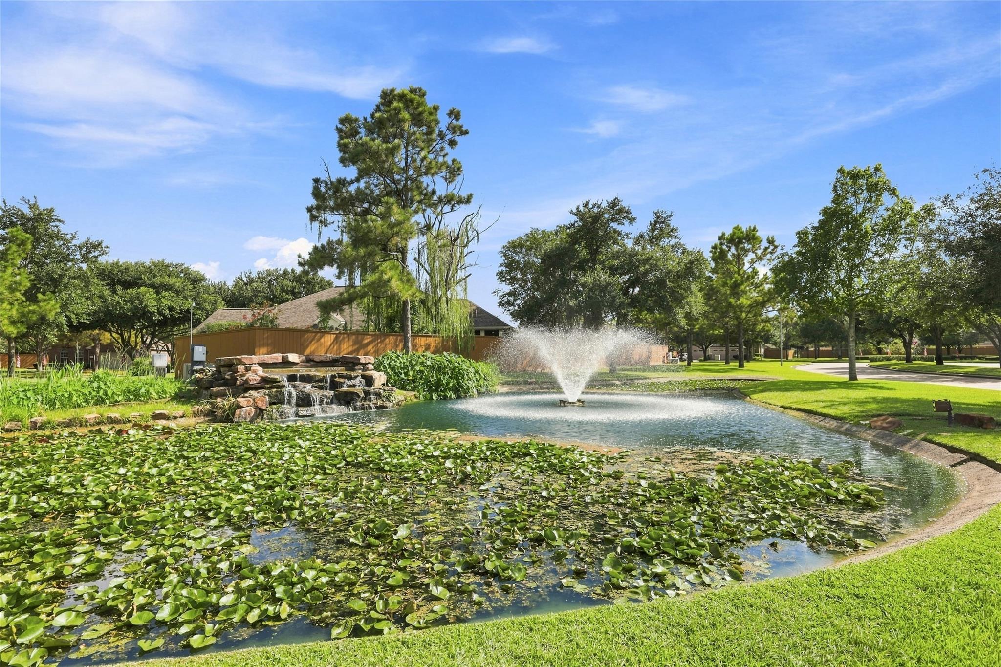 Community pond with a fountain, rock waterfall, and lily pads beside a grassy path.