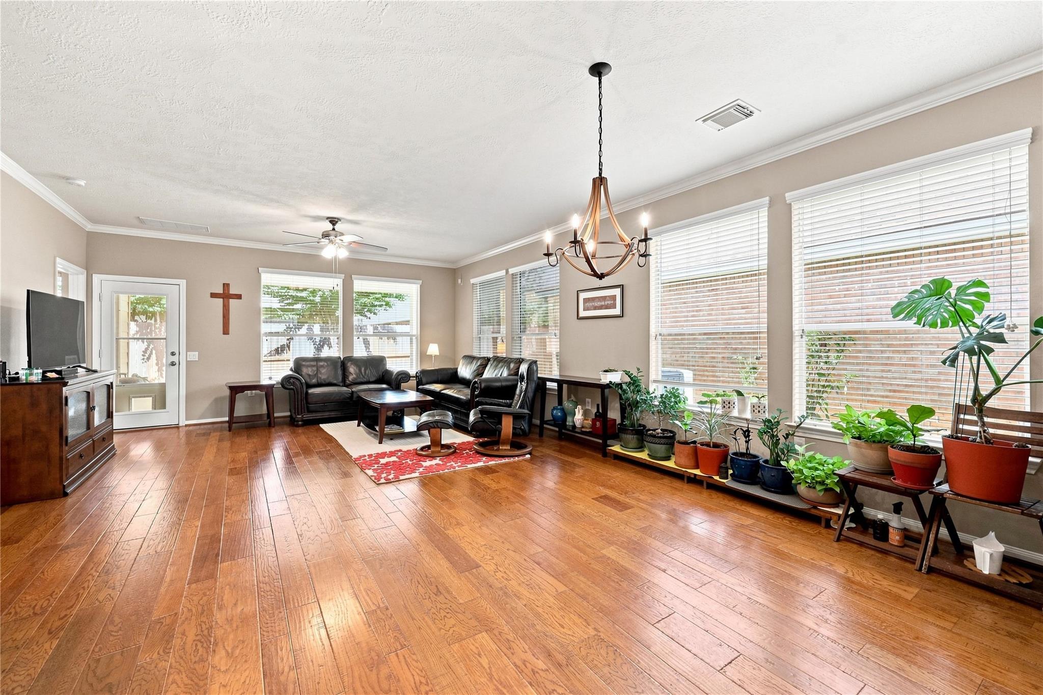 Living room with hardwood floors, leather seating, and potted plants beside large windows.