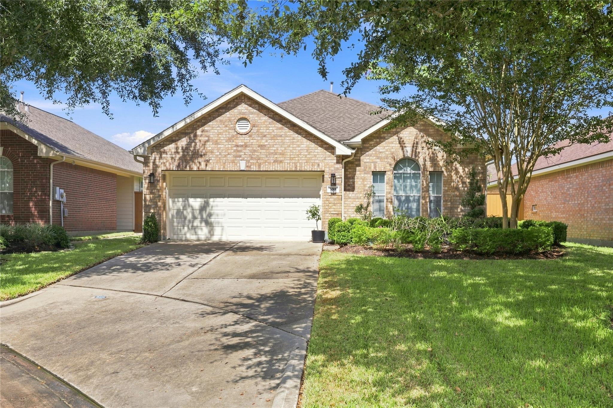 Tan brick home with a two-car garage, arched window, landscaped lawn, and wide driveway.