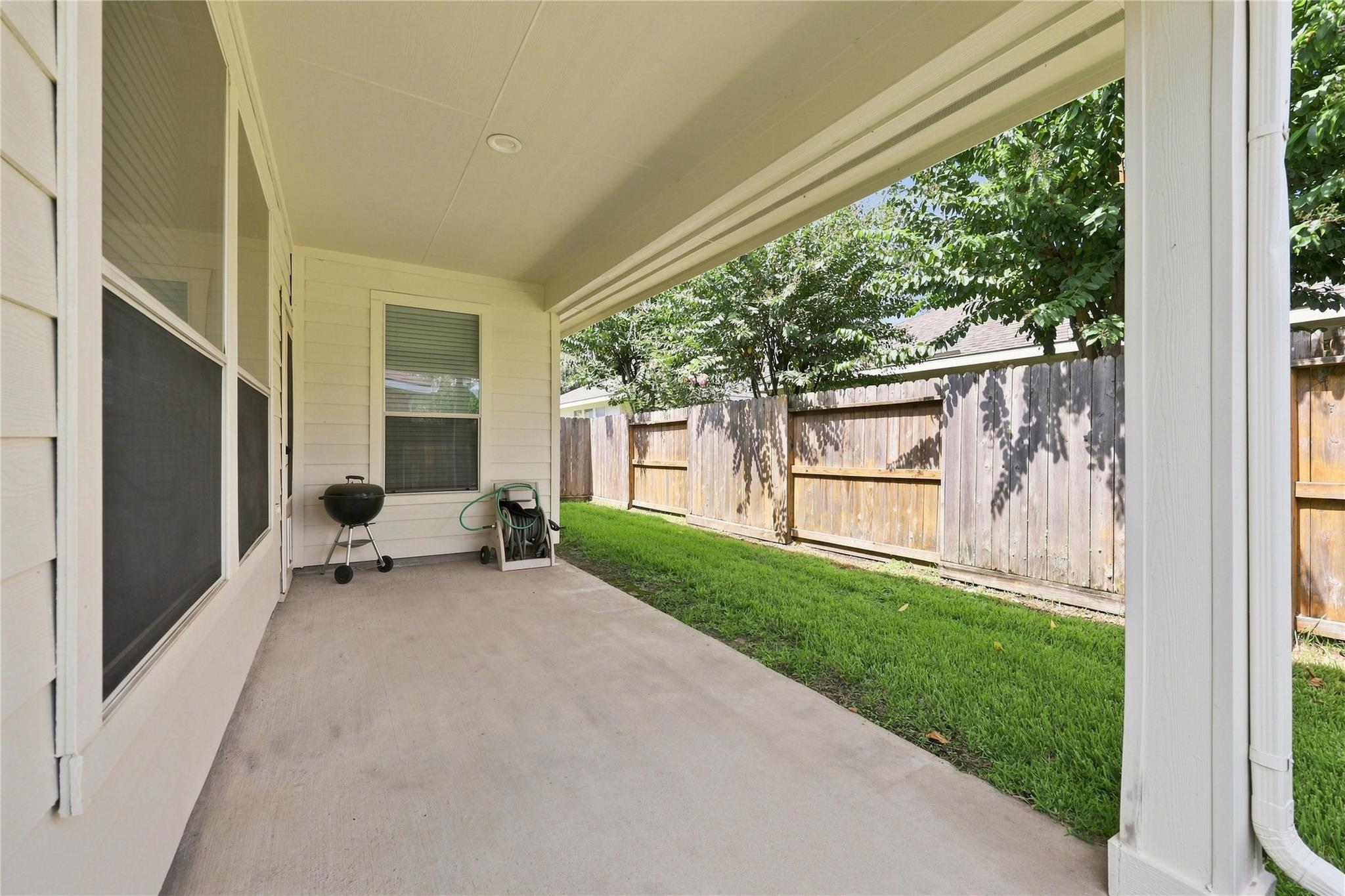 Covered patio beside a fenced grassy backyard, with a kettle grill and leafy trees.