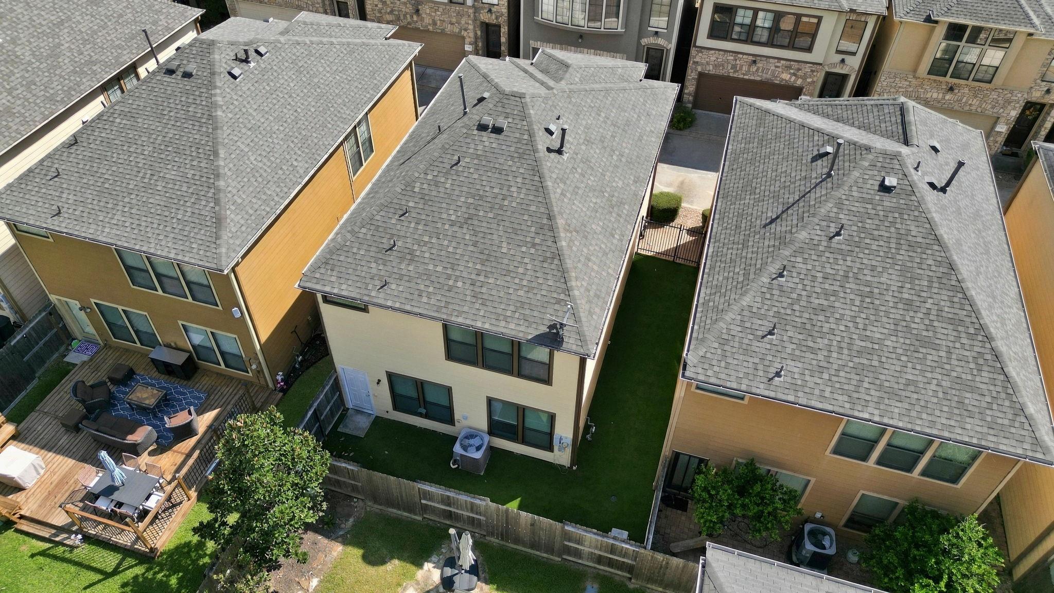 Aerial view of townhomes with gray shingle roofs and fenced backyards, including a furnished wooden patio.