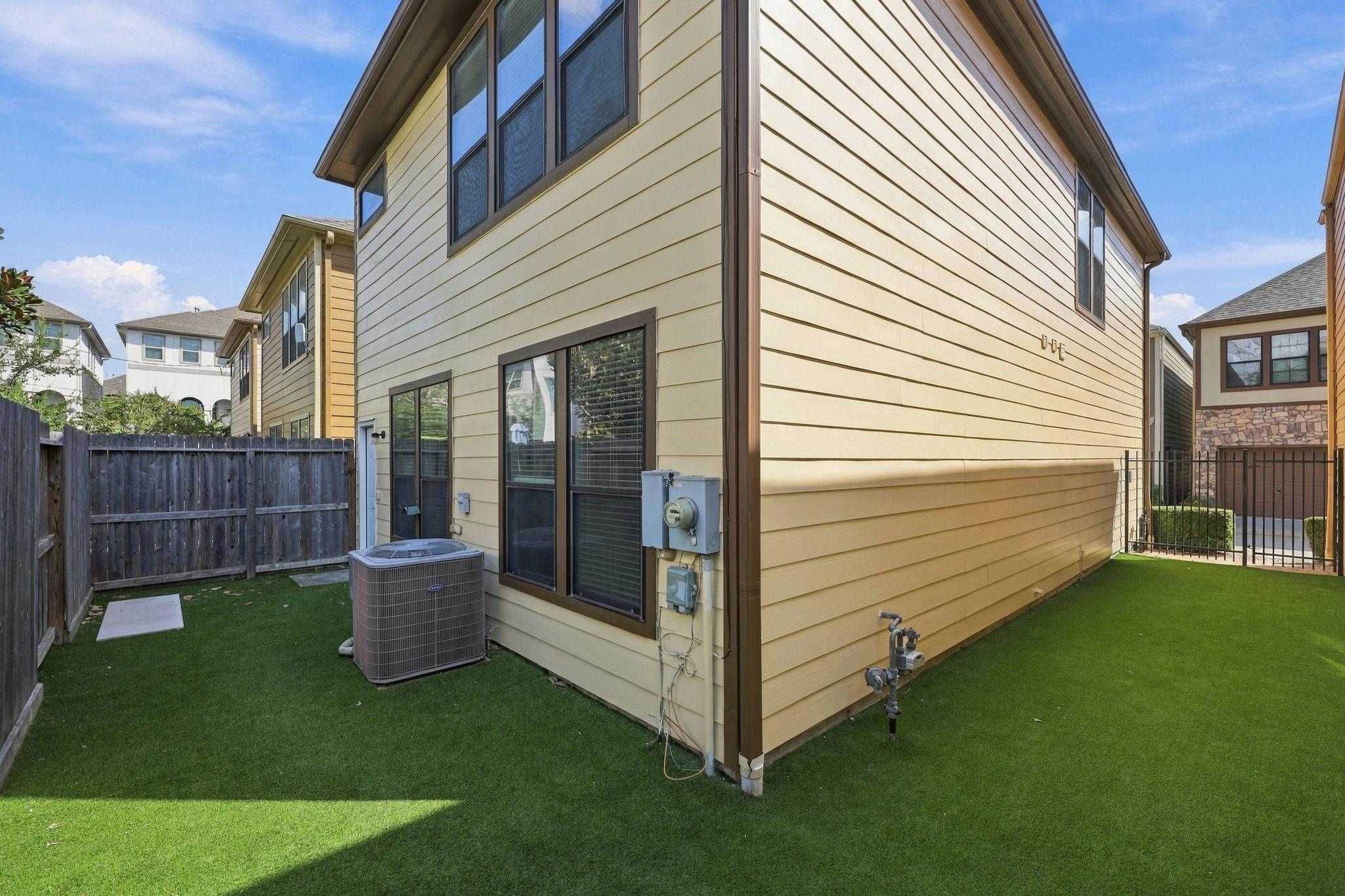 Two-story beige house beside a fenced artificial-turf yard, with dark-trimmed windows and an outdoor air-conditioning unit.