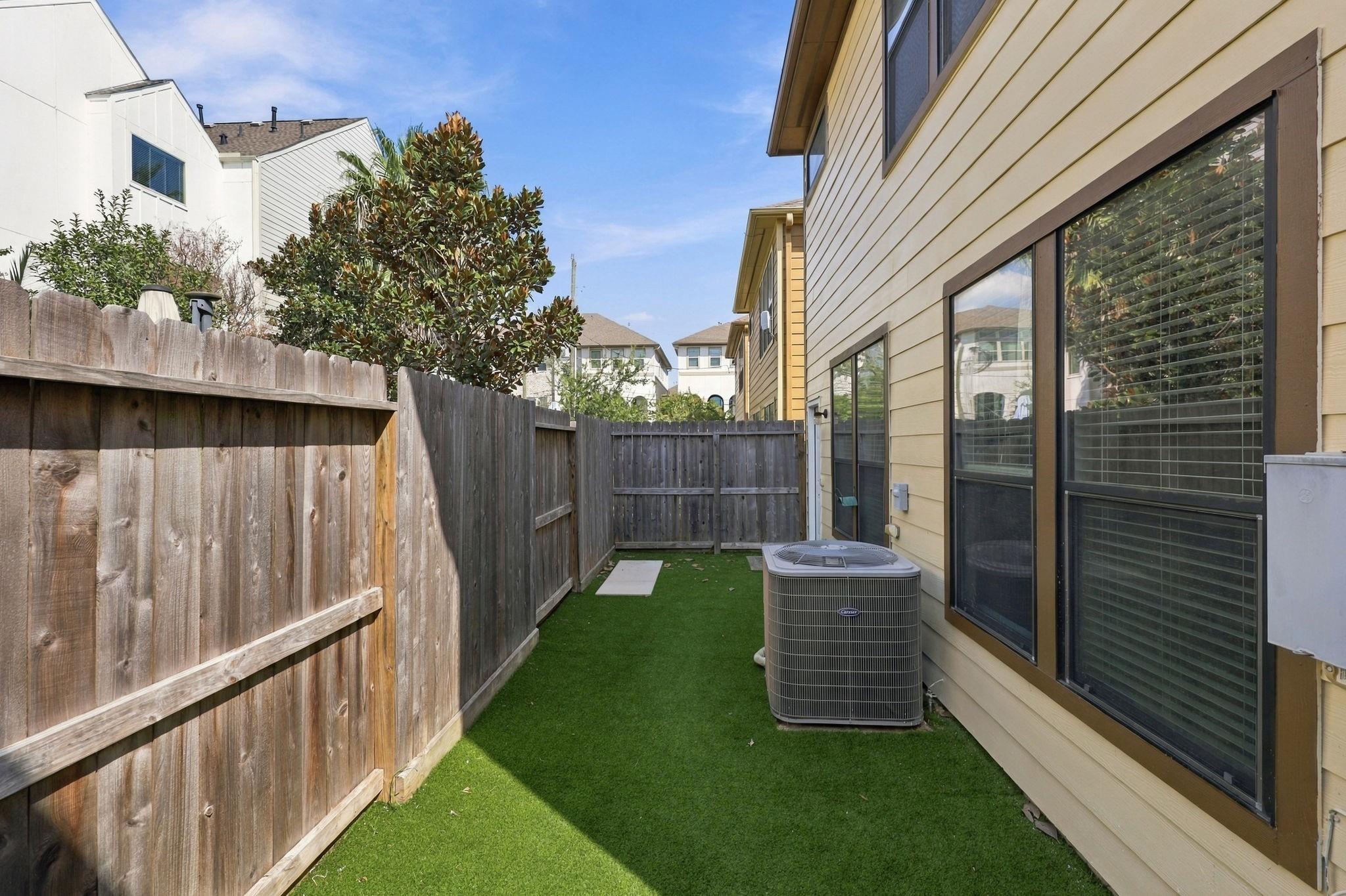 Fenced side yard with artificial turf, large windows, and an outdoor air-conditioning unit.