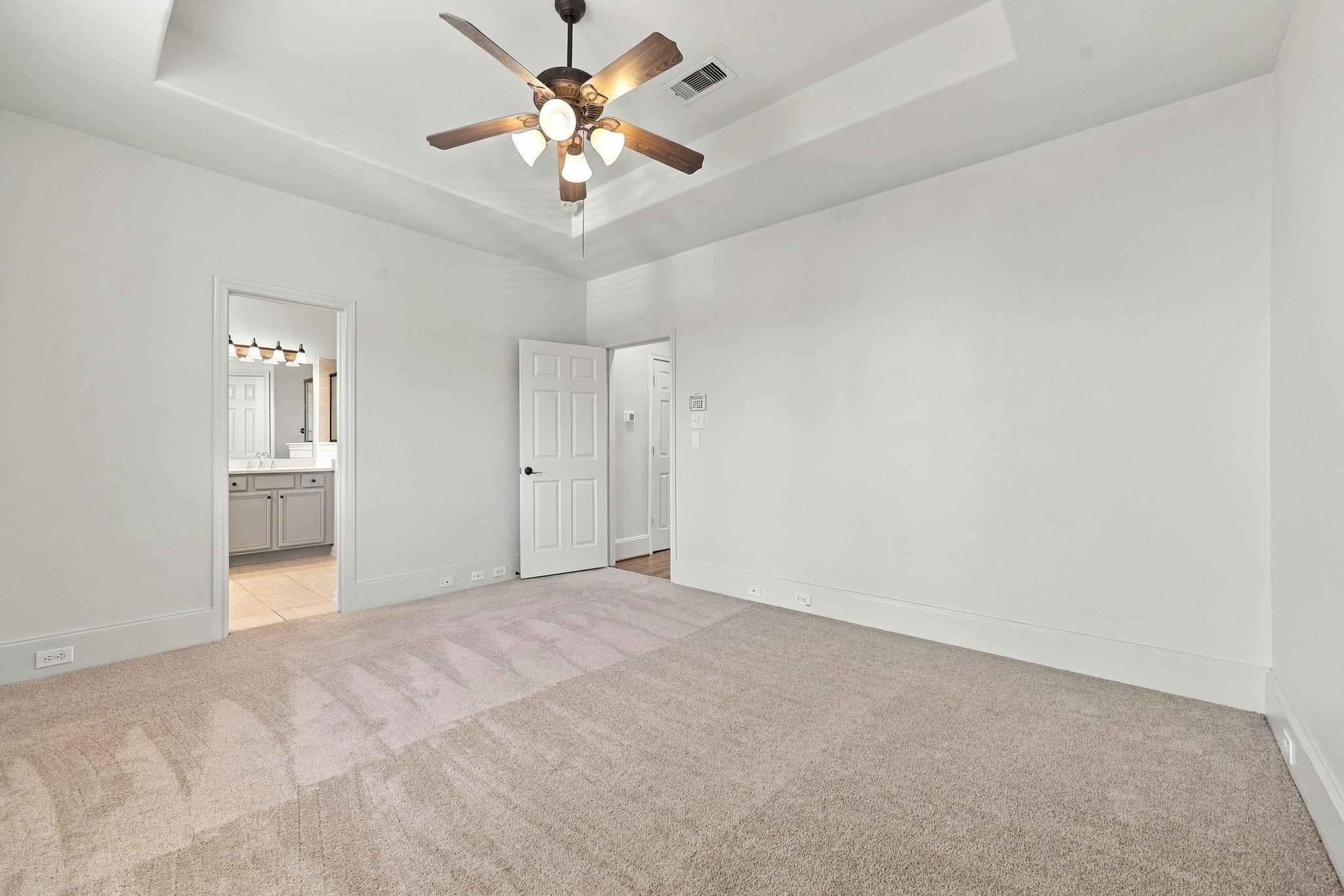 Empty carpeted bedroom with a tray ceiling and ceiling fan; bathroom vanity visible through doorway.