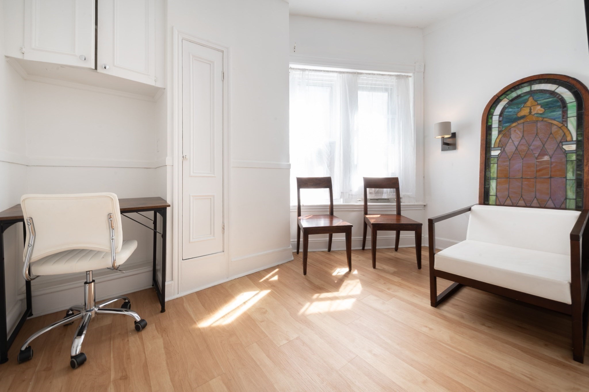 Bright sitting room with hardwood floors, a desk, and a settee before a stained-glass panel.